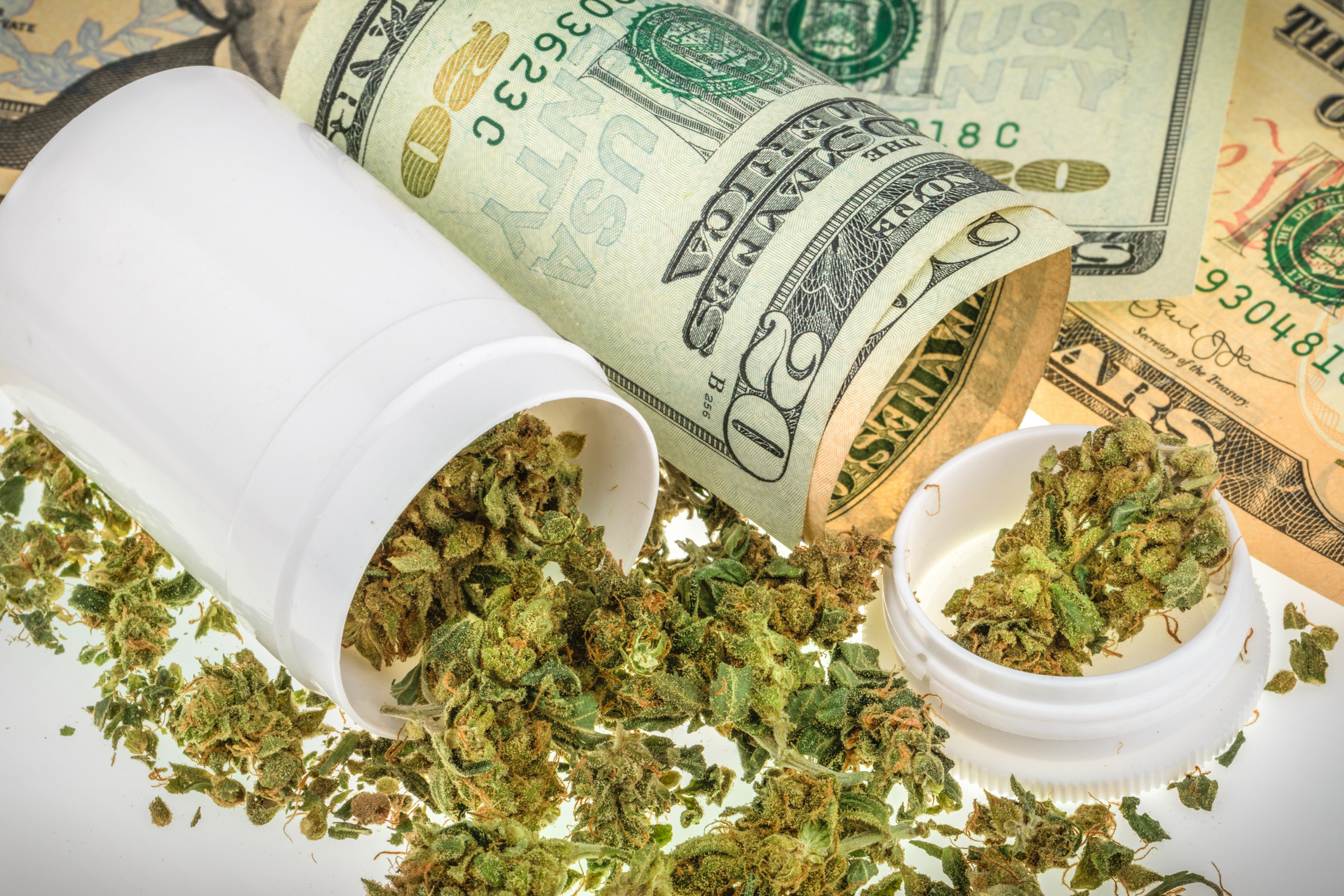 A tipped over bottle filled with dried cannabis buds sitting next to a messy pile of cash bills.