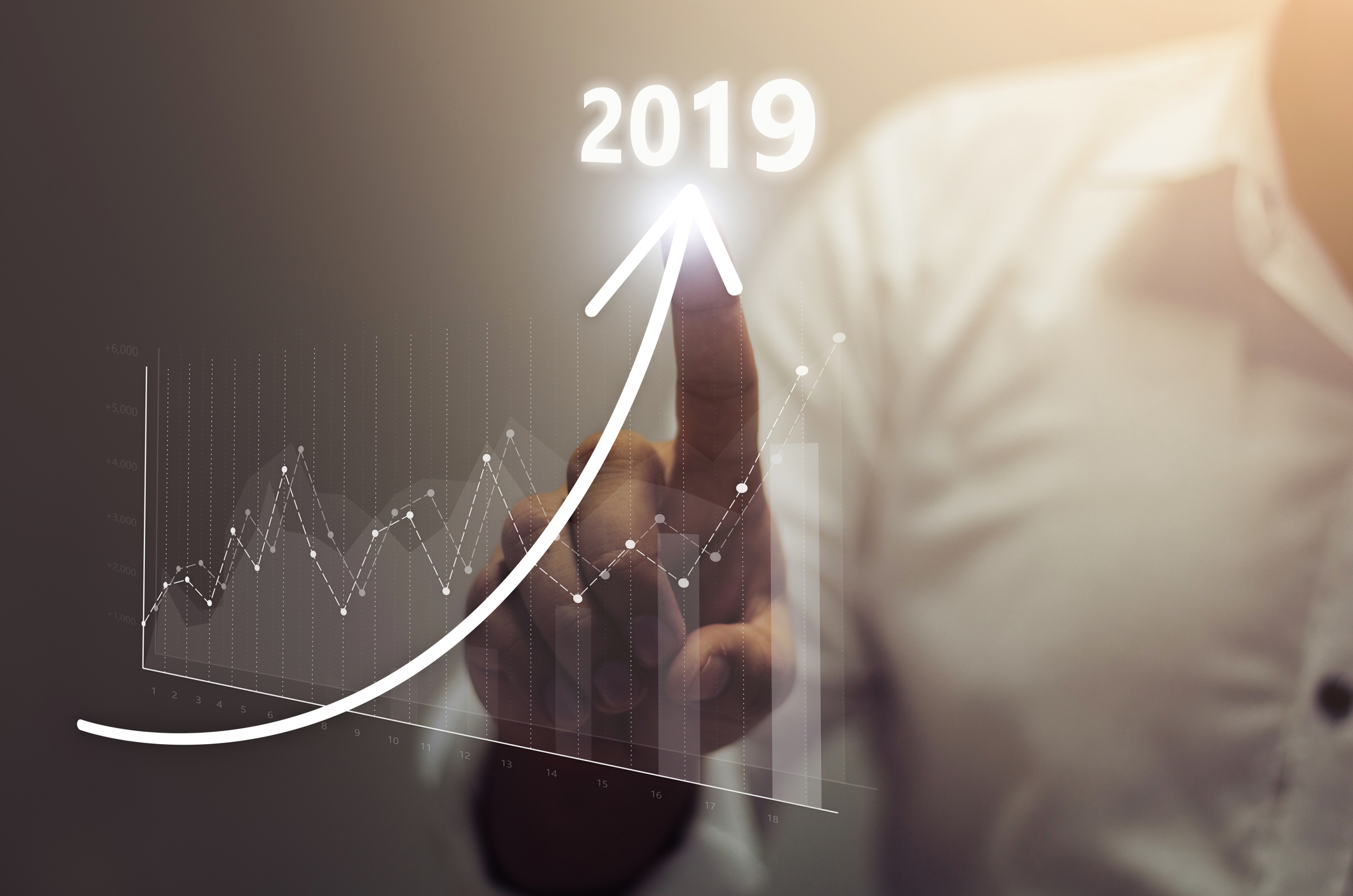 A businessmans finger tracing an upward arrow labeled 2019, with a graph showing uneven but significant growth.