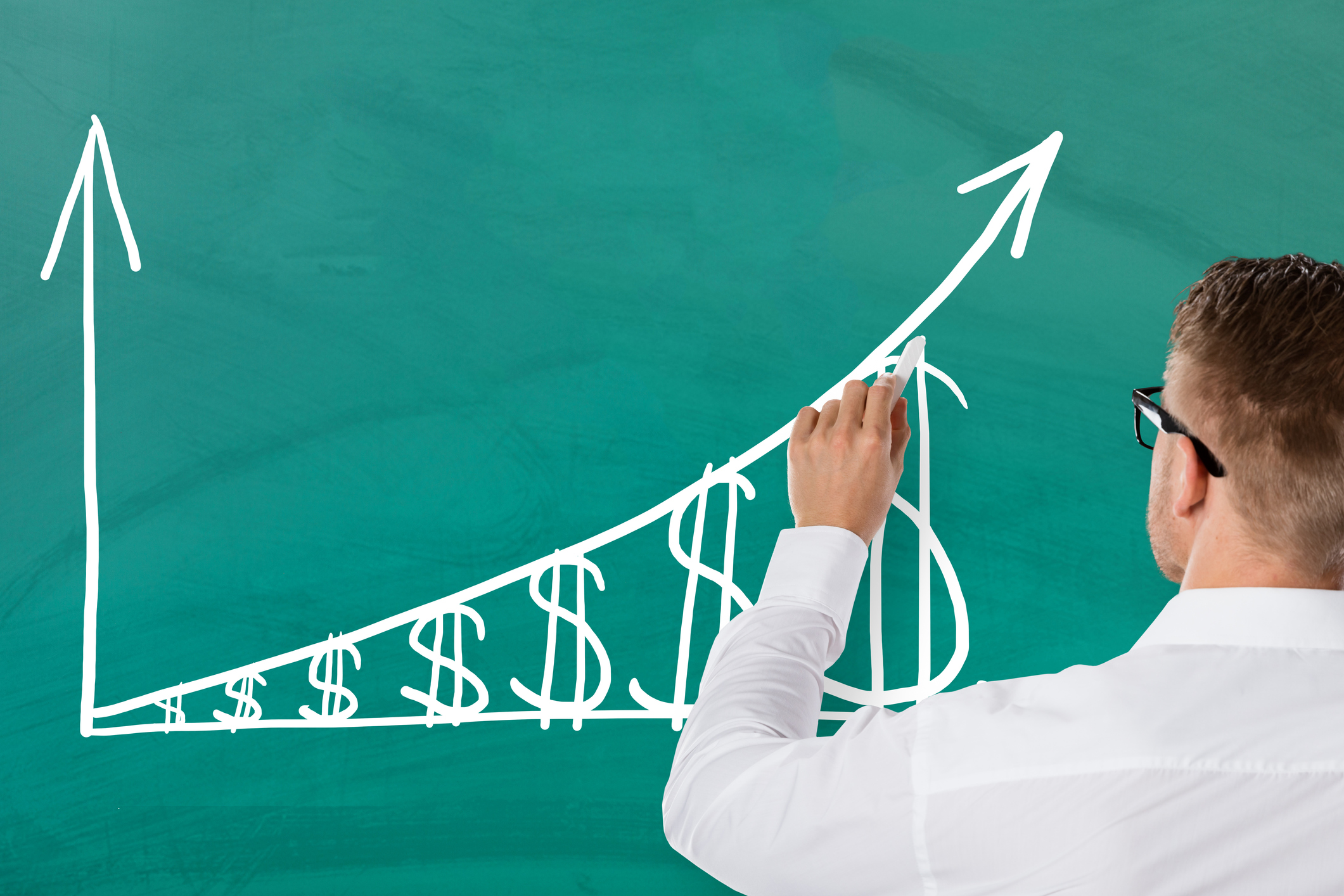 A person drawing a chart on a green chalkboard of rising dollar signs.