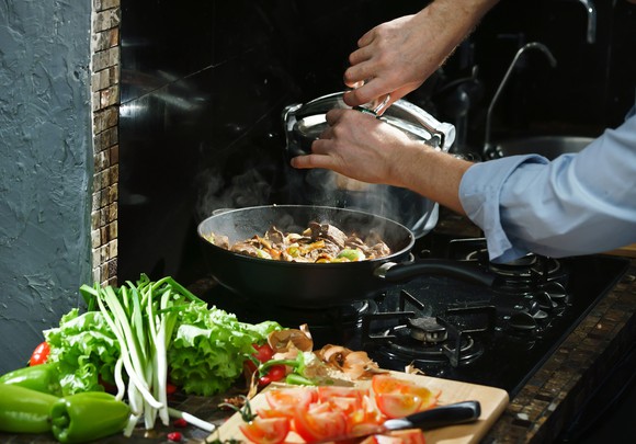 Two hands pouring spices into a frying pan on a stove, with veggies on the side counter.