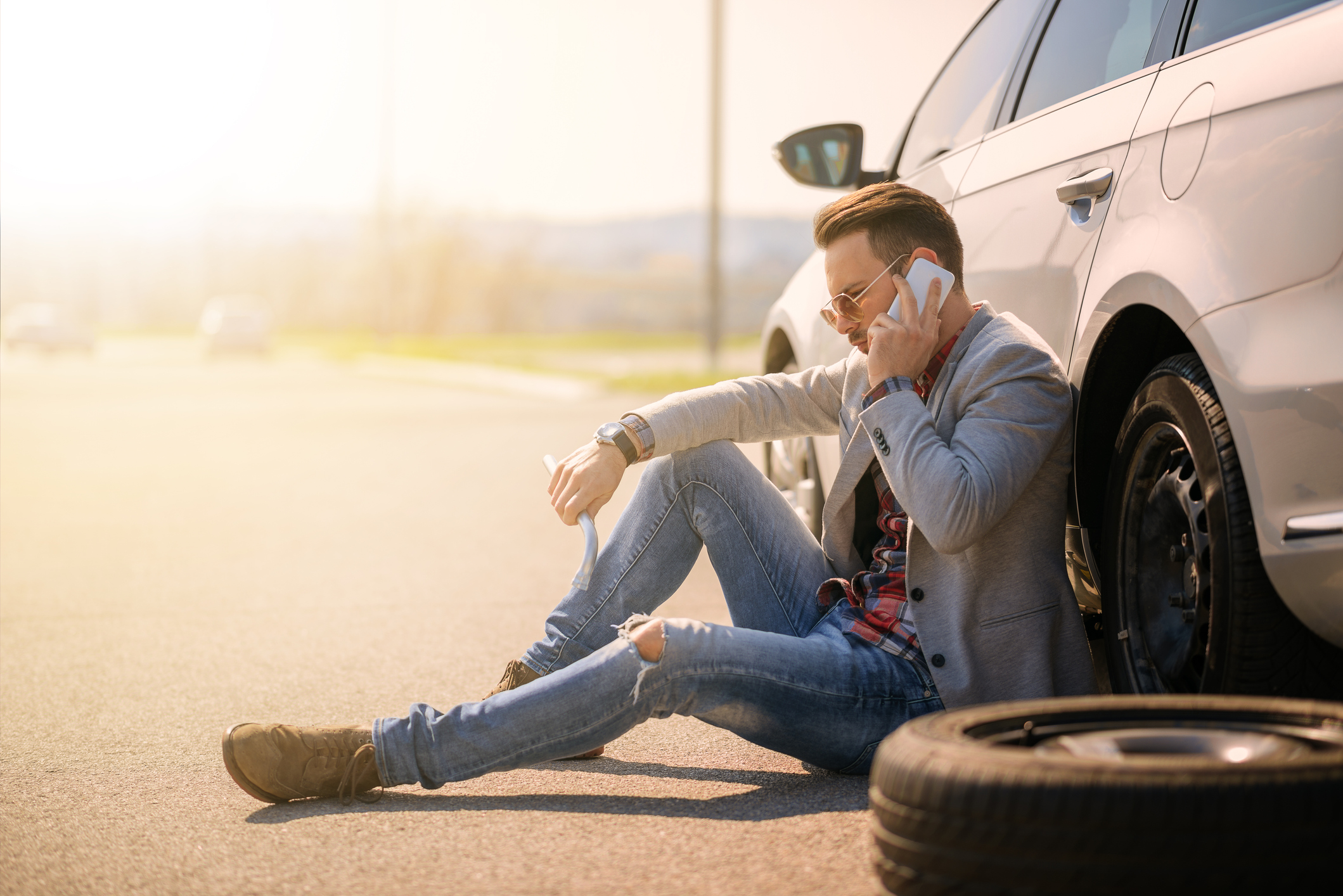 Man sitting against car on the side of the road, talking on a smartphone