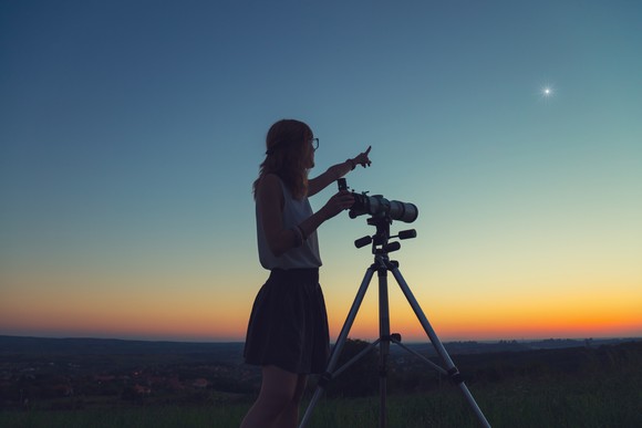 A woman stands next to a telescope at dusk and points to a bright star int he evening sky. 