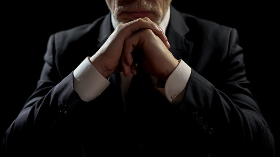A man in a suit viewed from the neck down rests his chin on folded hands which rest on a table below.