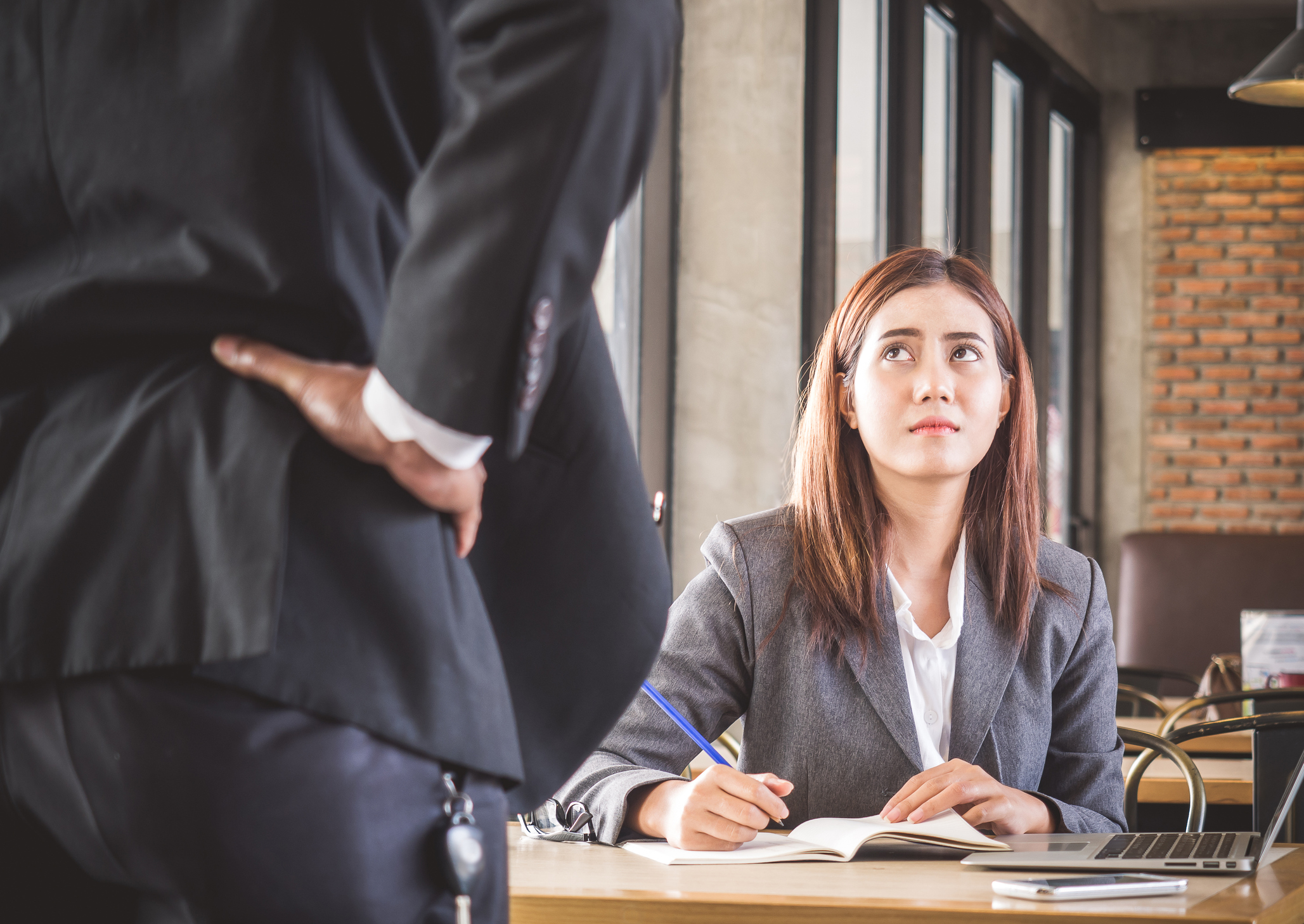 Woman in business suit seated at desk with annoyed expression looks up at man in suit who has his hands on his hips.