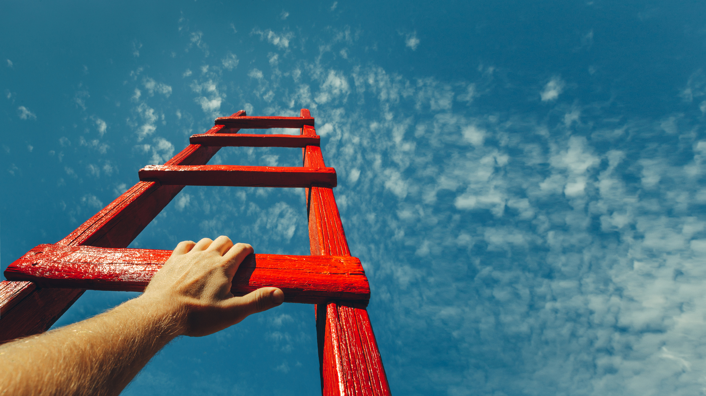 A hand holding the bottom rung of a red ladder set against a clear, blue sky.