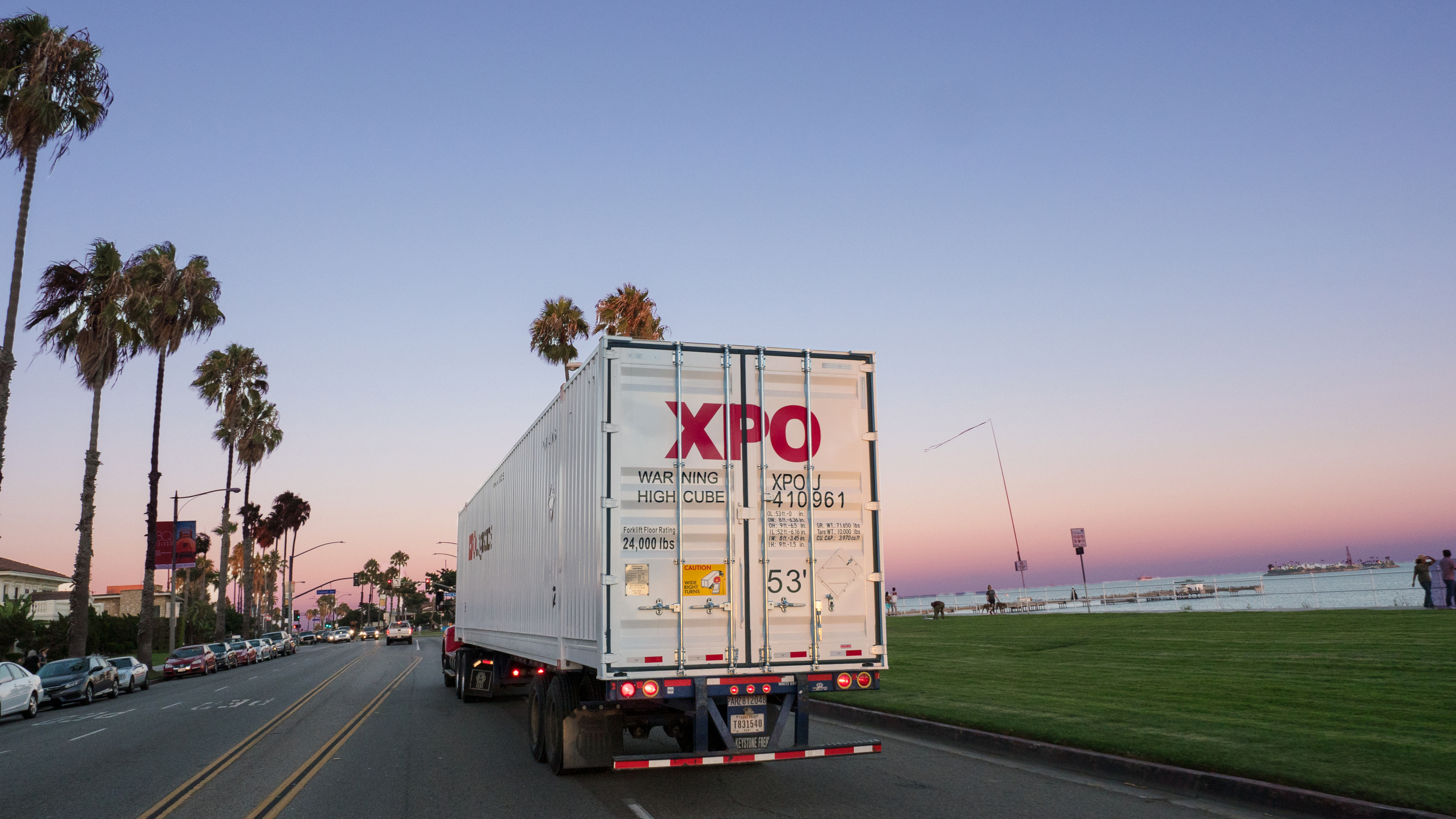 An XPO truck on the road near water, surrounded by palm trees and blue sky
