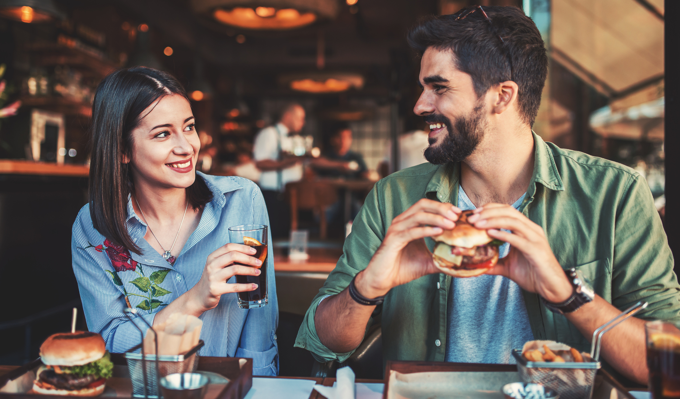 A young man and woman sit side by side smiling at each other in a restaurant. The man holds a burger, and the woman holds a drink.