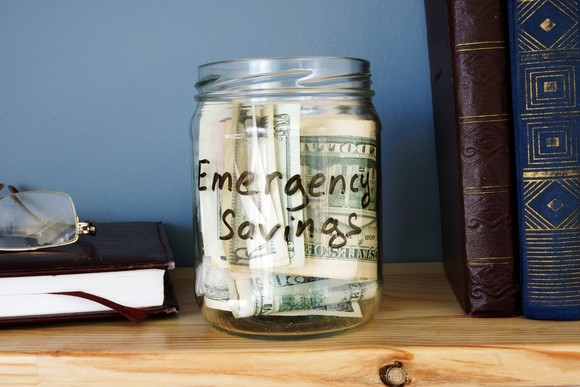A glass jar filled with cash and labeled emergency savings sits on a wooden shelf, between books and a pair of reading glasses.