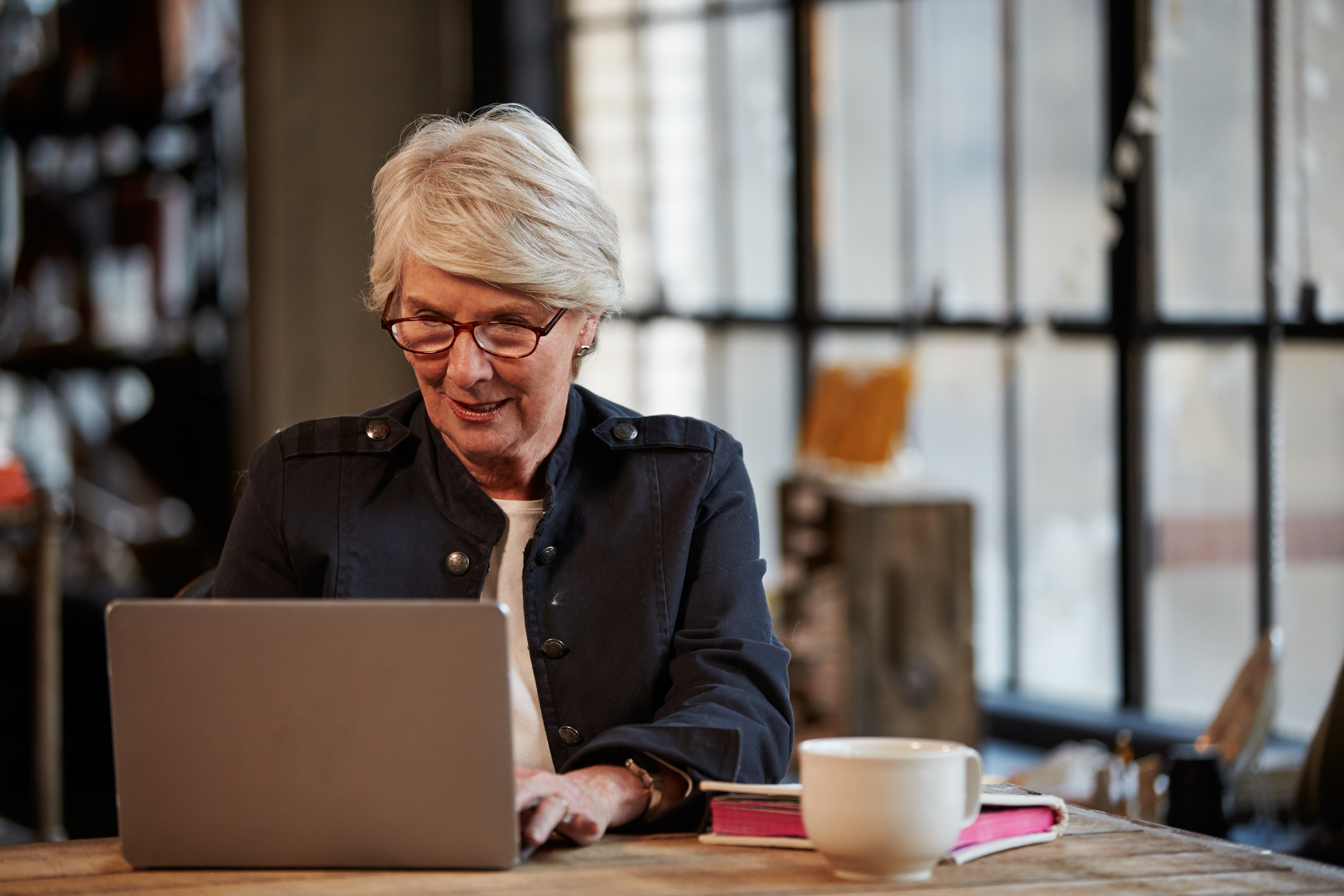 Older woman typing on laptop; next to laptop are a mug and book