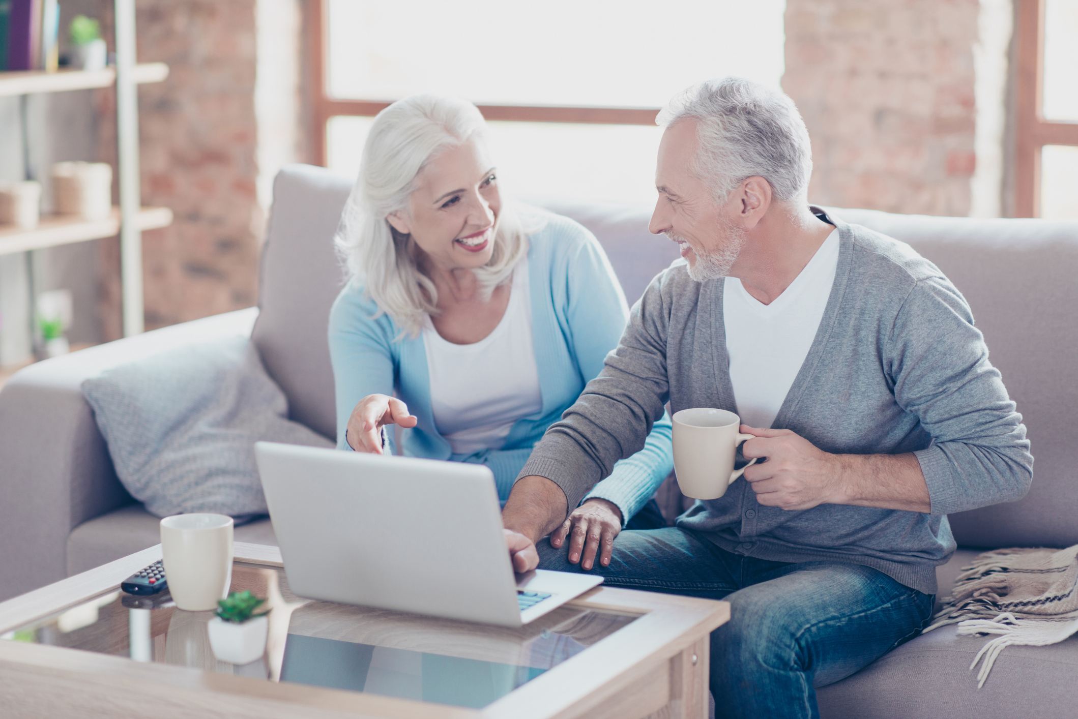 Smiling senior man and woman sitting on couch, facing each other while man's hand hovers over laptop on coffee table