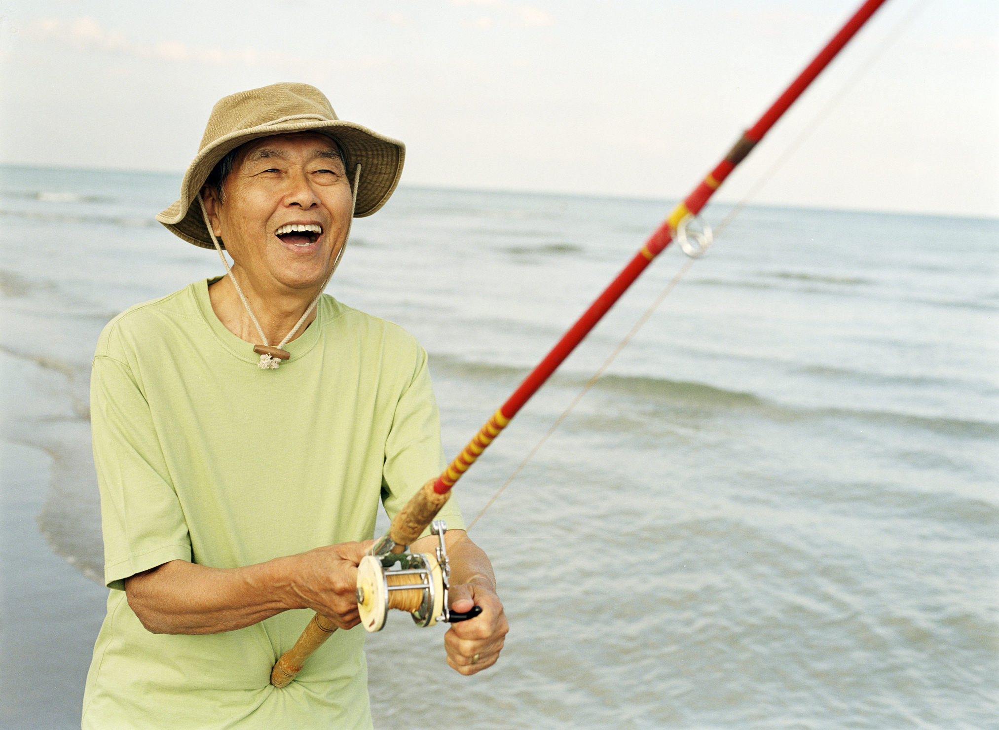Laughing older man holding red fishing pole, wading in the waves.
