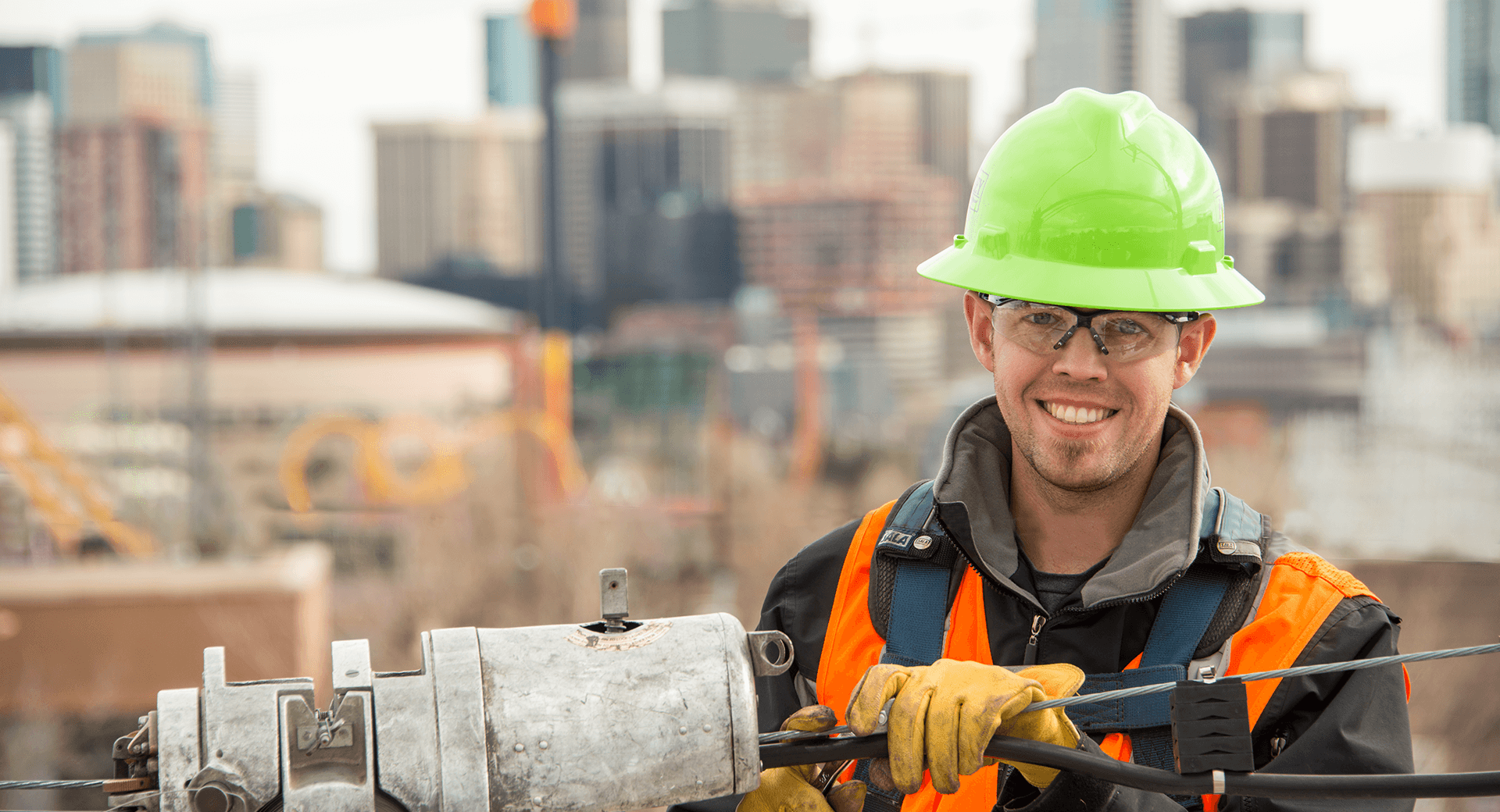 Smiling worker with green hard hat connecting telephone wire in front of a city skyline.