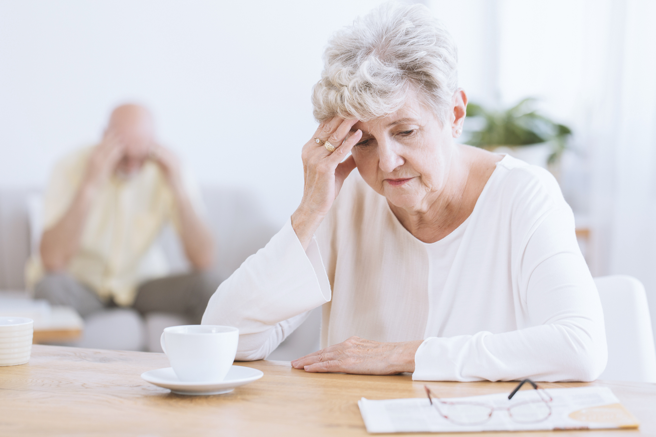 Older woman seated at table, holding her head with serious expression