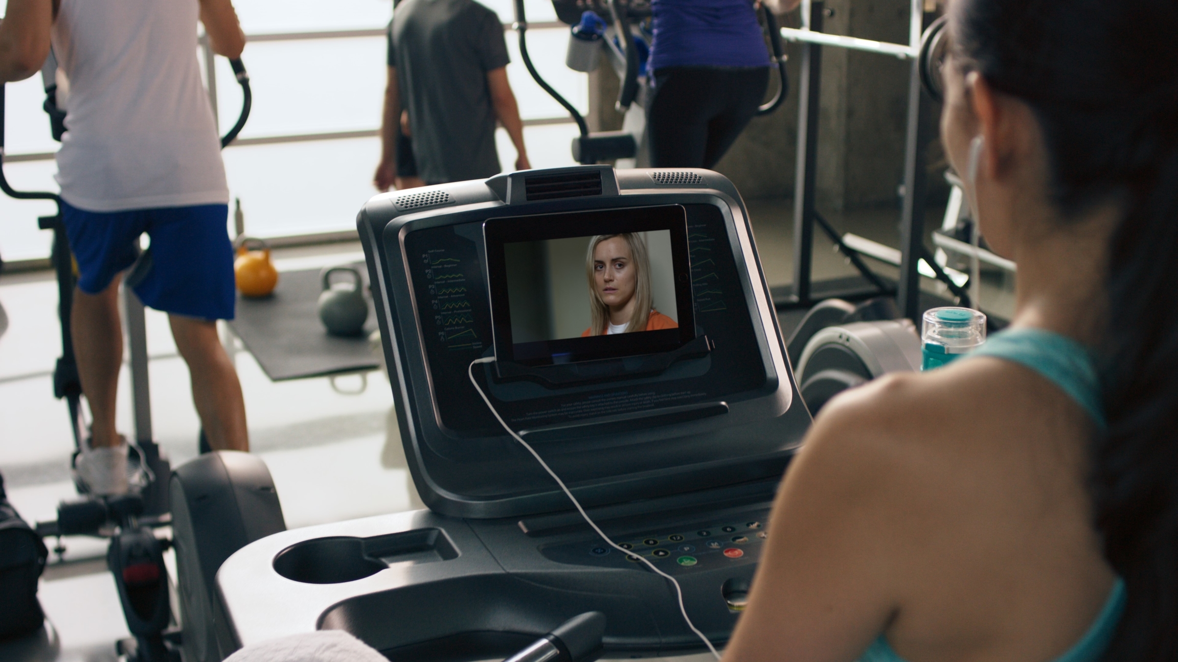 A woman on an exercise machine watching Netflix original series "Orange Is the New Black" on a tablet while she exercises in a crowded gym.