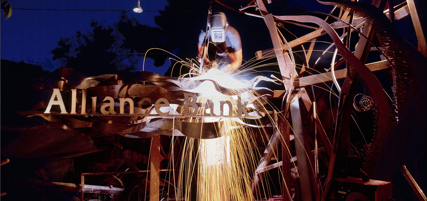 Welder working on metal sculpture with Alliance Bank logo in it.