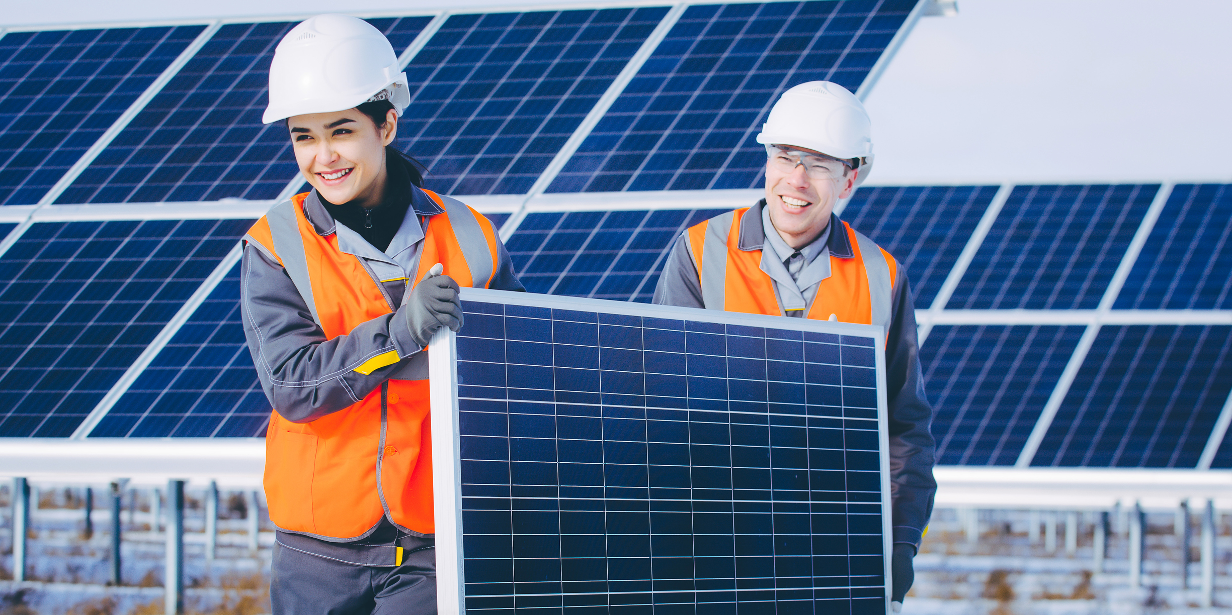 Two people carrying a solar panel with a solar farm in the background.