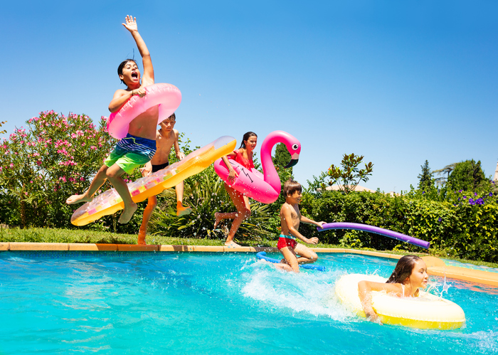 Three young boys and two young girls -- each with an inner tube or raft -- in various states of jumping into an in-ground swimming pool with foliage and blue sky in background.