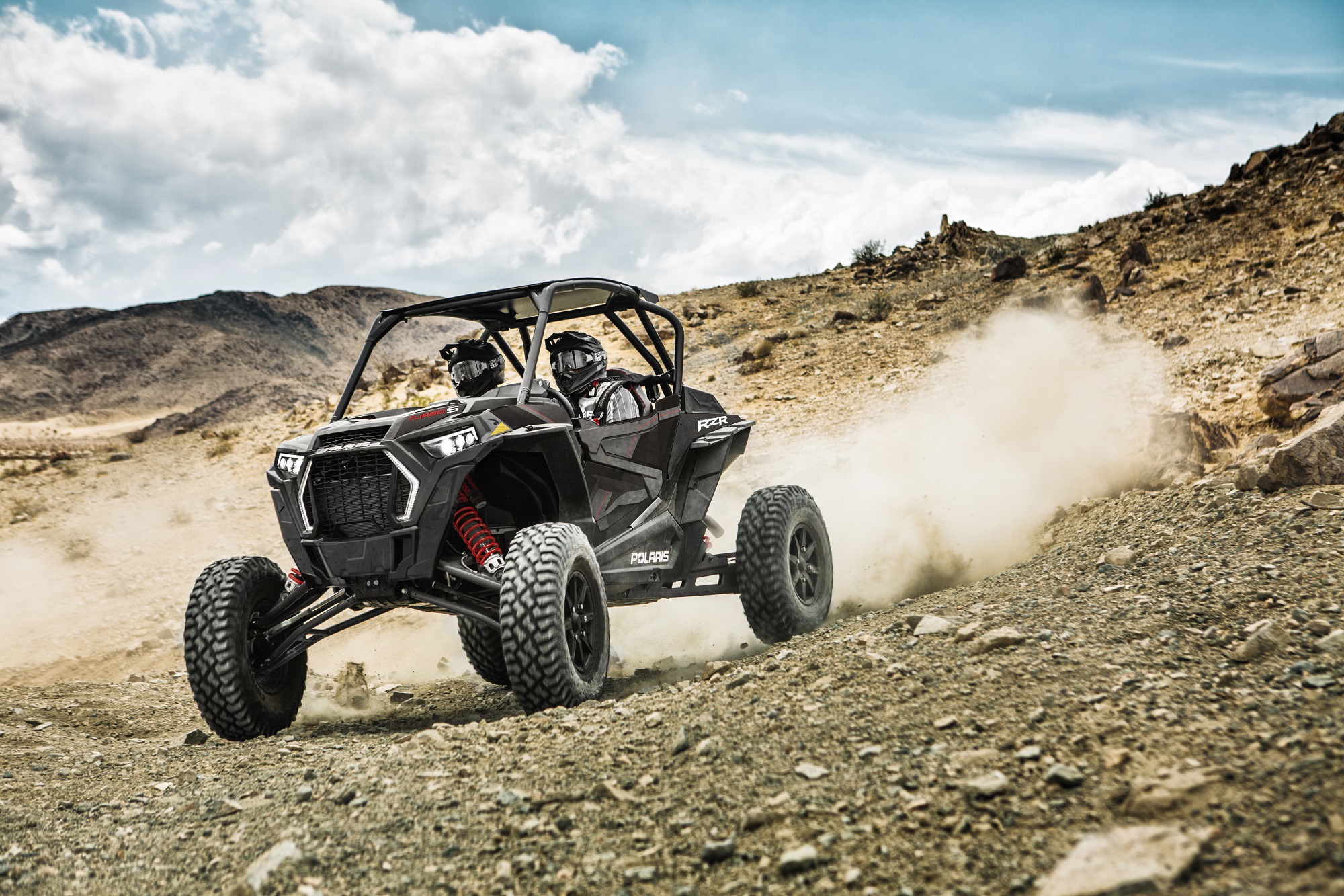 Two-seat Polaris RZR spraying dust behind it on a rocky mountain landscape.