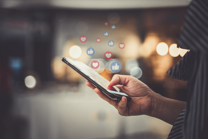 Person's hand holding a cell phone with common social media icons (for like and love) floating above the phone.
