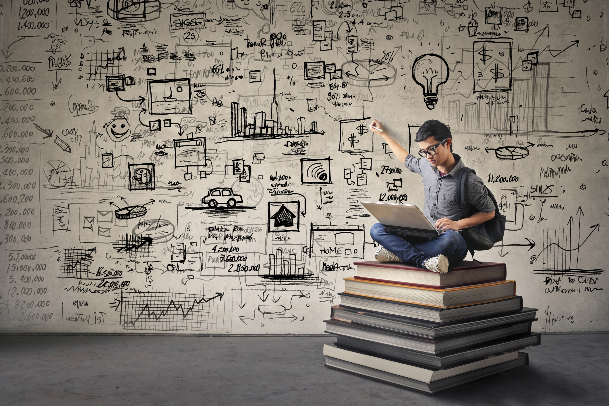 A man sitting on a stack of books while pointing at diagrams written on the wall behind him.