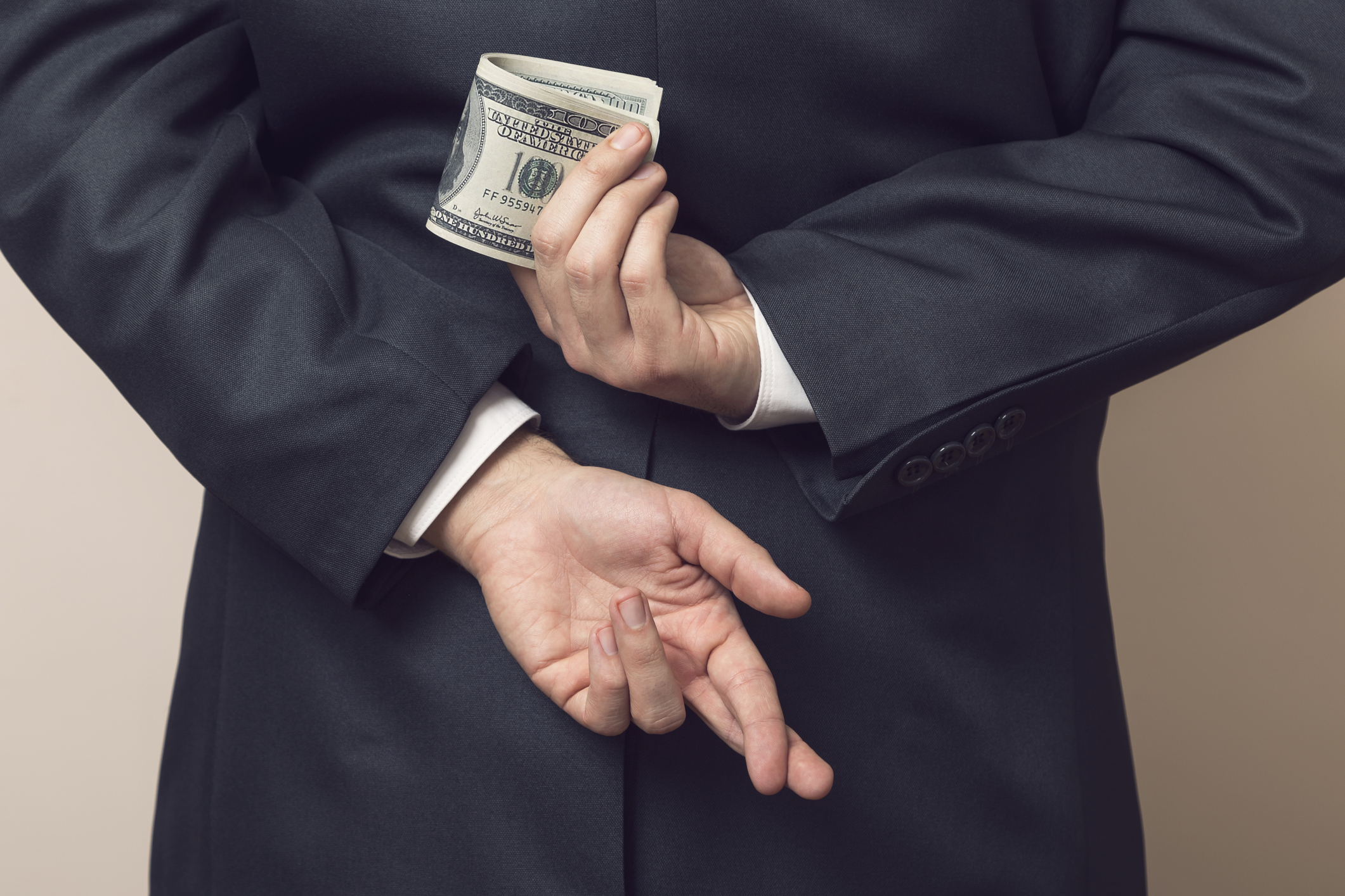 A businessman crosses his fingers behind his back while holding a wad of hundred dollar bills.