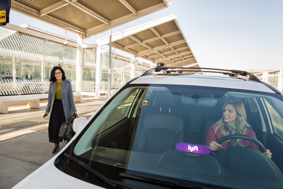 A woman in the driver's seat of a parked car with the Lyft logo on the dashboard while another woman approaches the car.