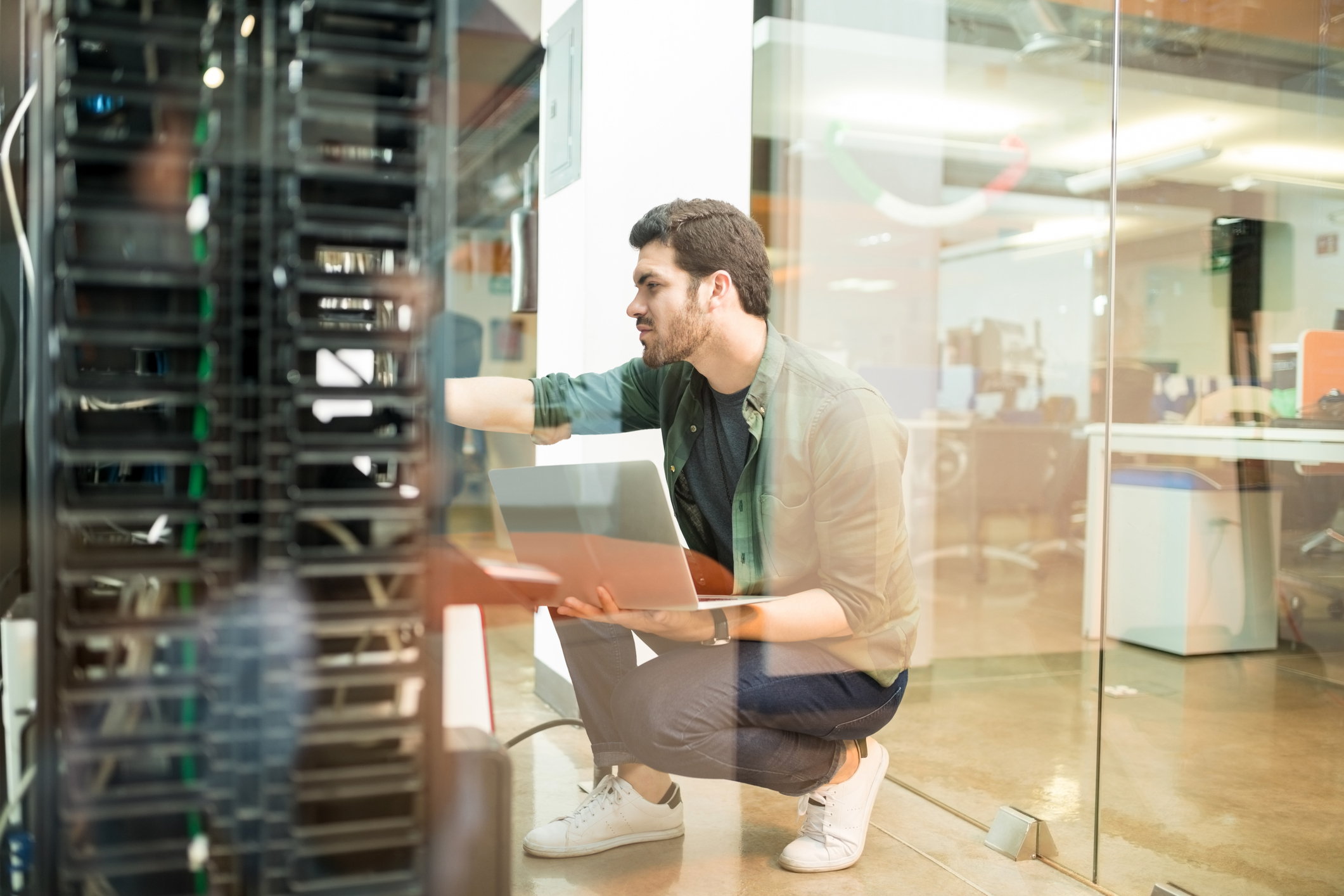 A young man holds a laptop while kneeling and checking out a rack of servers in an IT room.