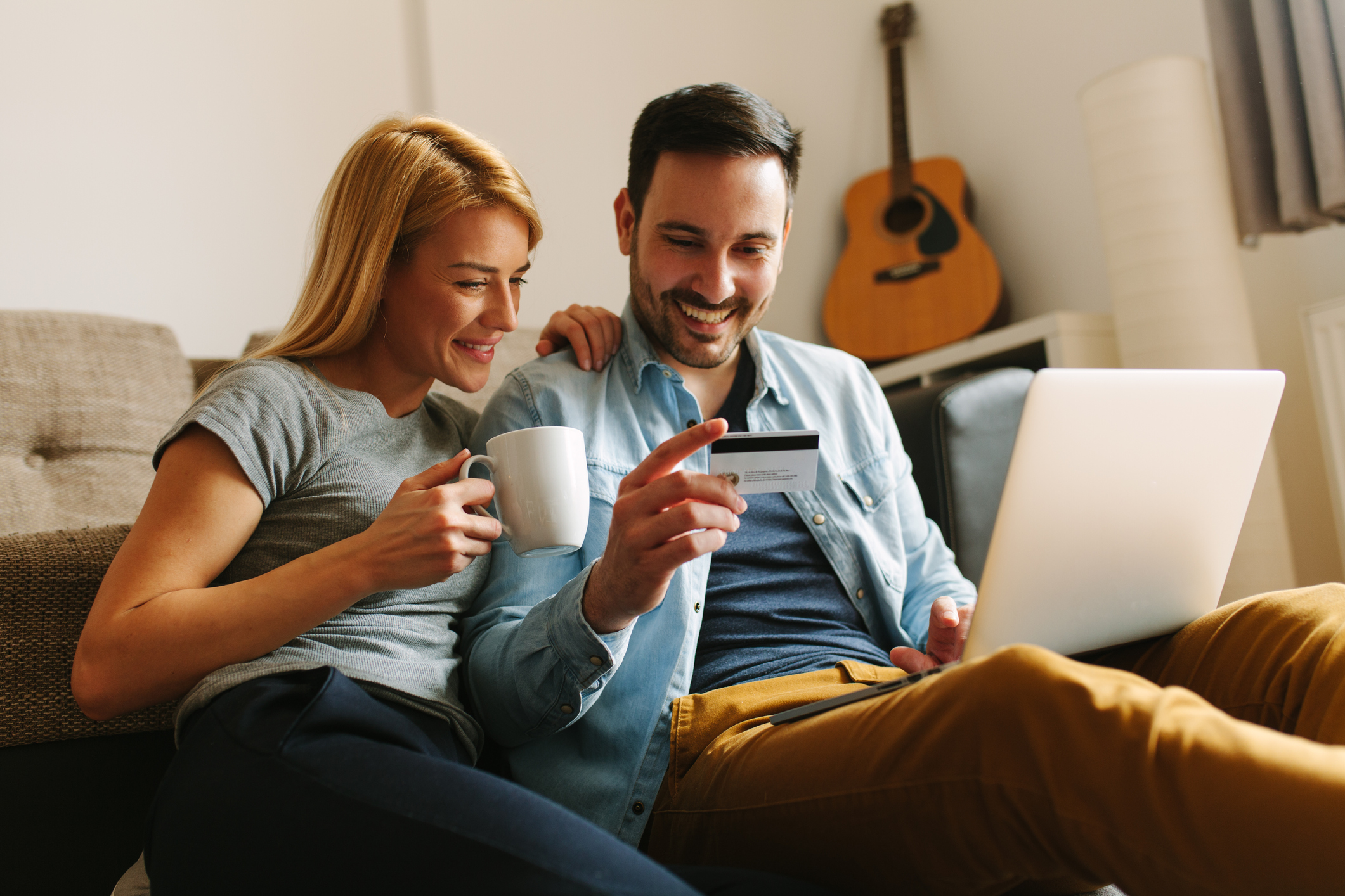 A woman holds a cup of coffee, her hand on the shoulder of a man who's got his laptop open and a credit card in one hand.
