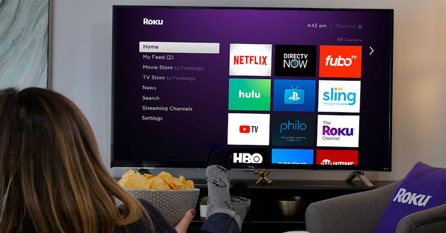 A woman, with her feet up on a coffee table, holding a bowl of chips while browsing the Roku homescreen on a TV.