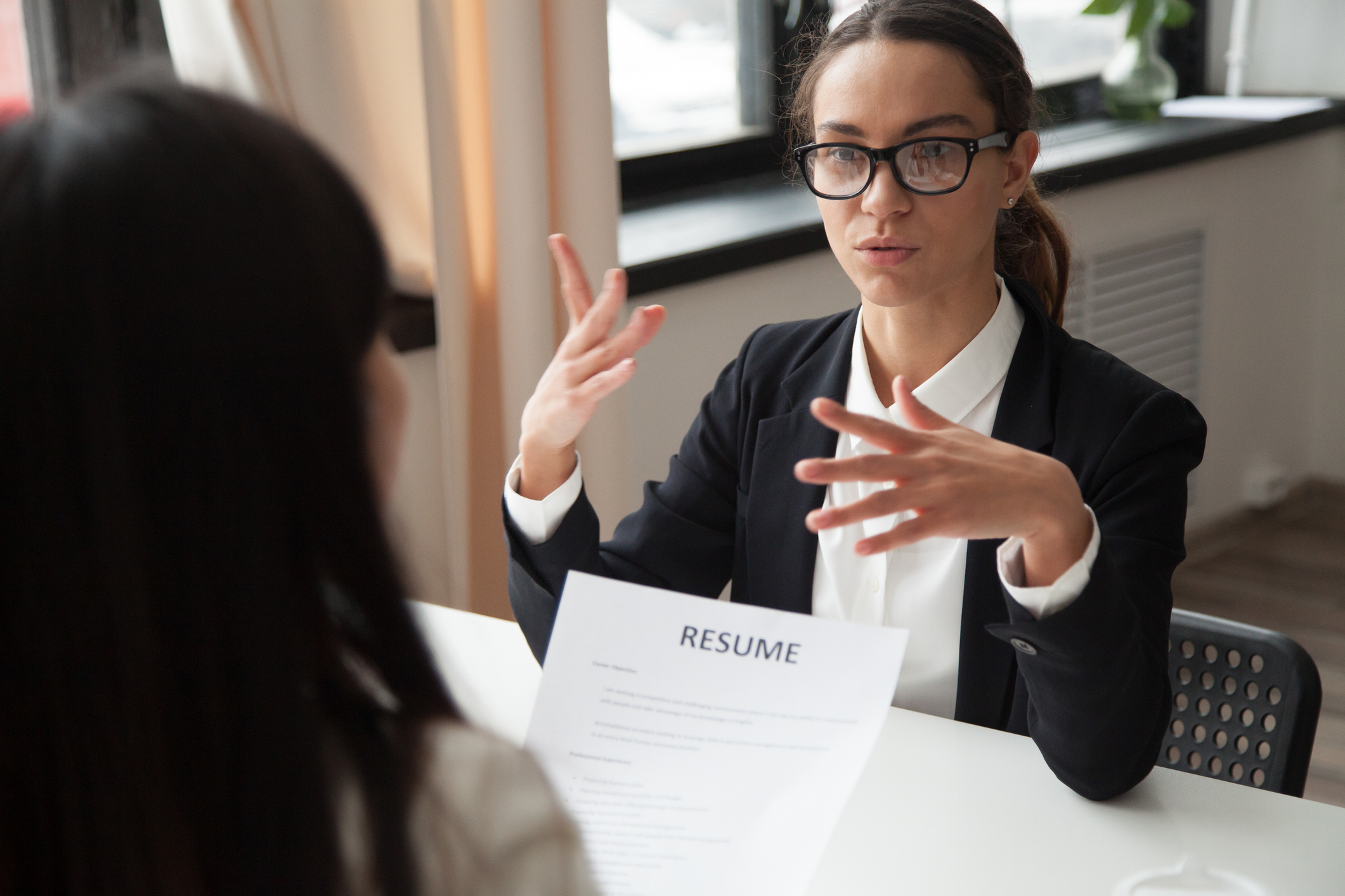 Professionally dressed woman gesturing while sitting across a table from another woman holding a resume