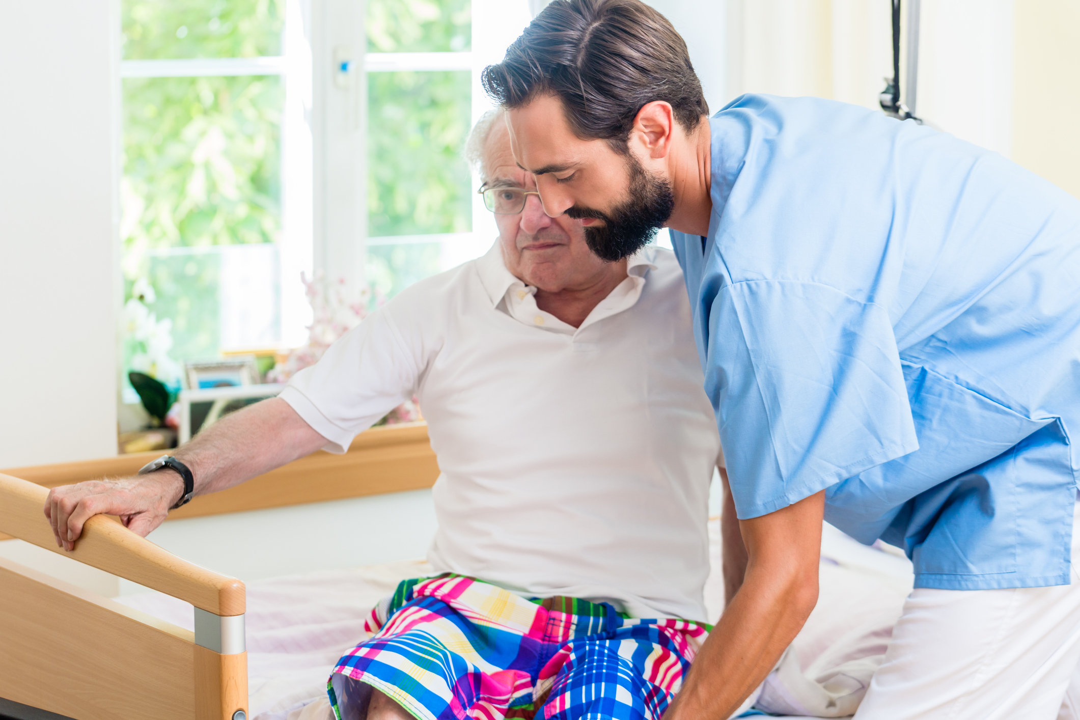 Man in scrubs helping older man into bed.
