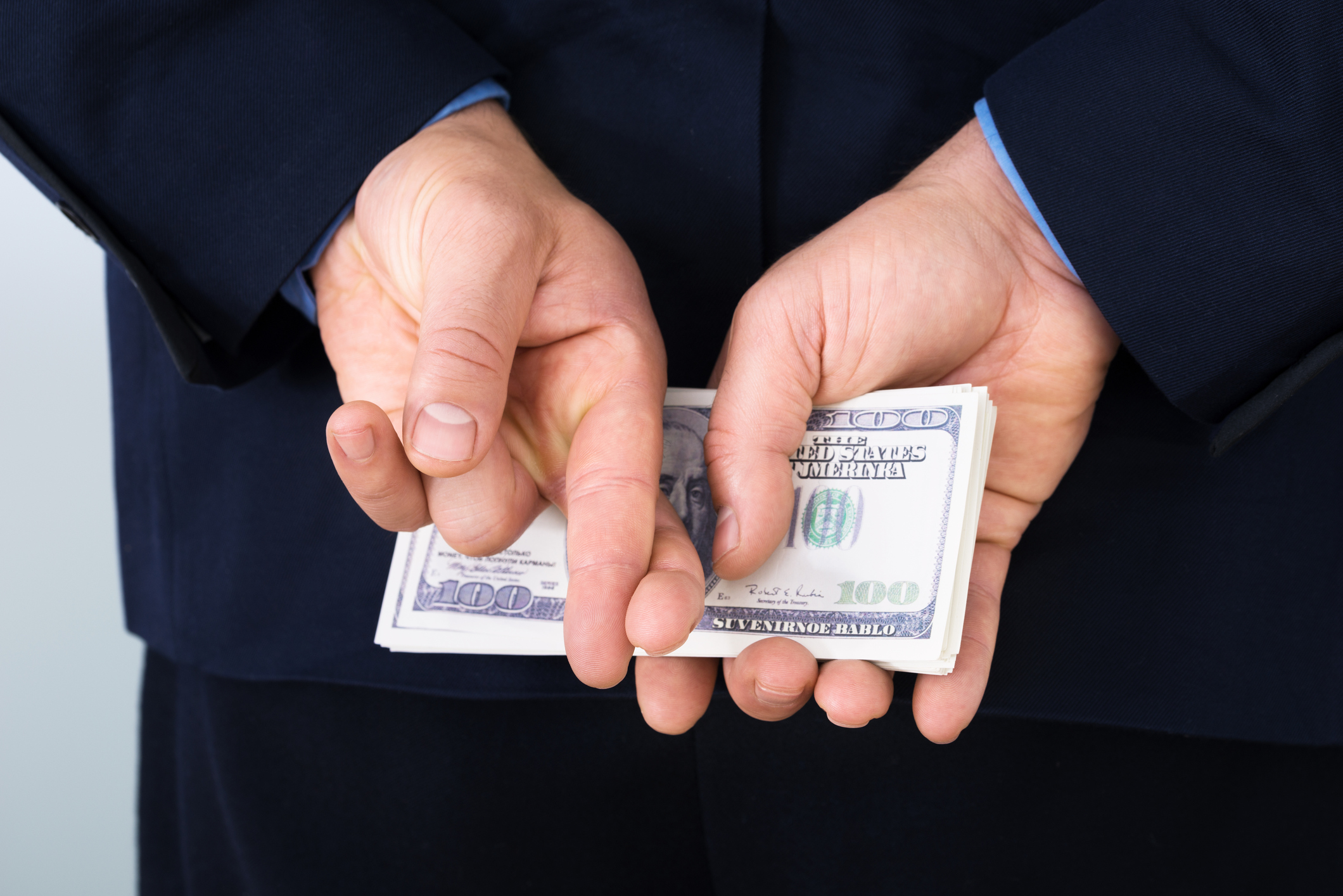 A man in a suit holding a stack of cash behind his back with his fingers crossed.