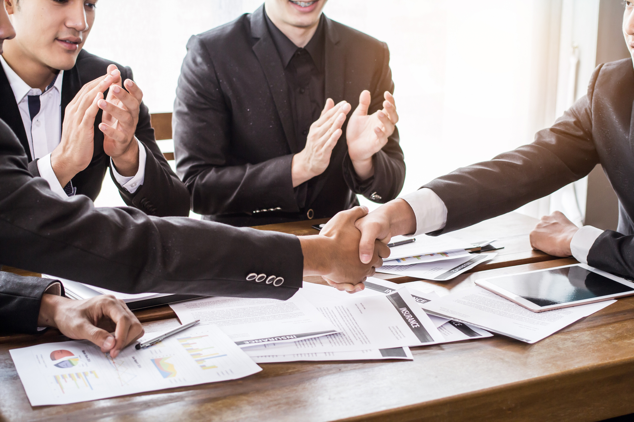 Two men shaking hands while others applaud at a table with papers spread out.