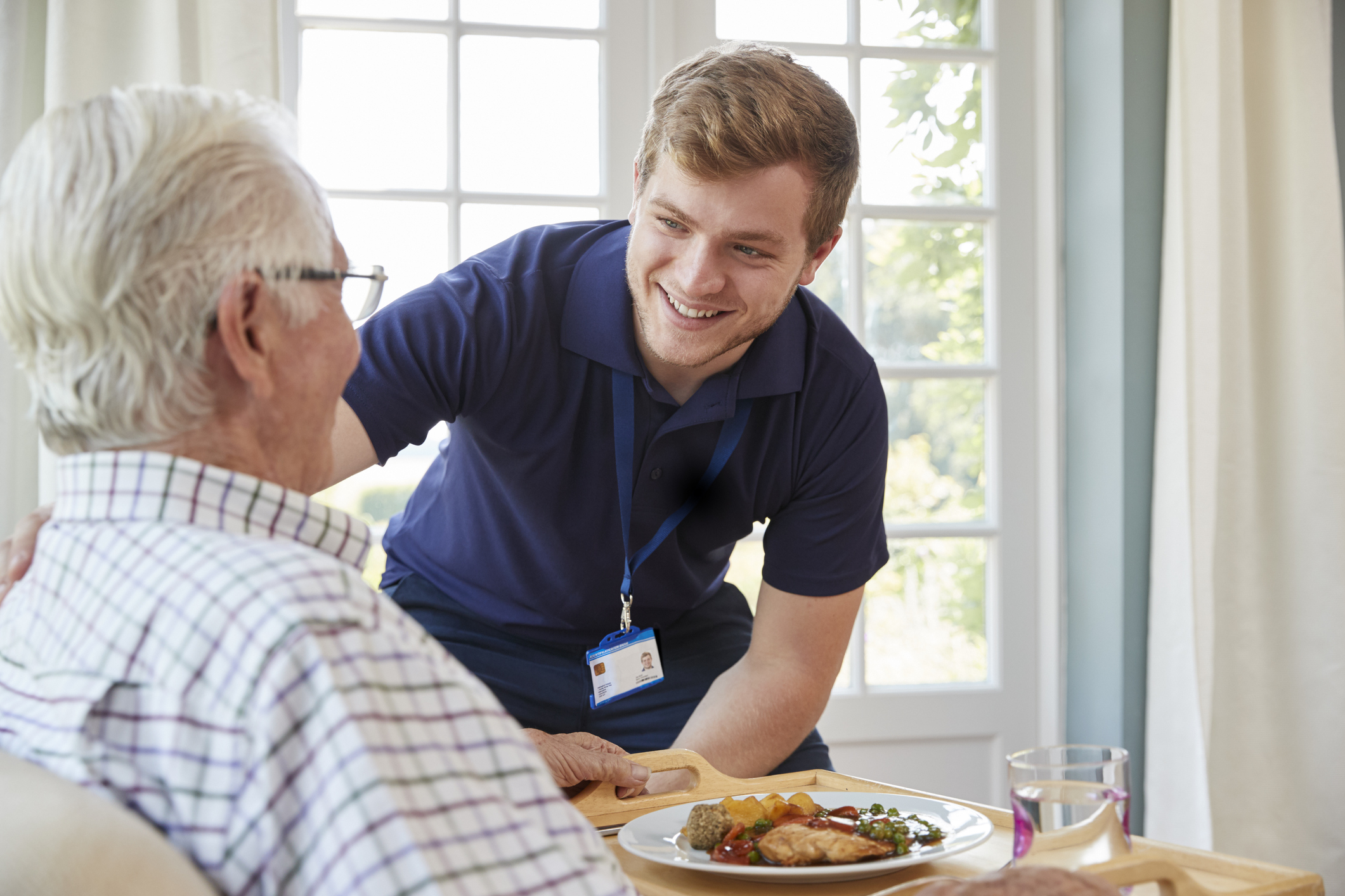 Young man in scrubs leaning over toward older man with meal in front of him