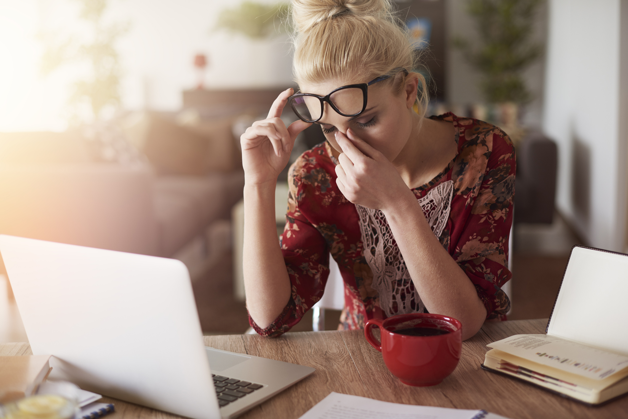 Woman sitting at a desk with an open laptop, holding her glasses and pinching her nose as if tired