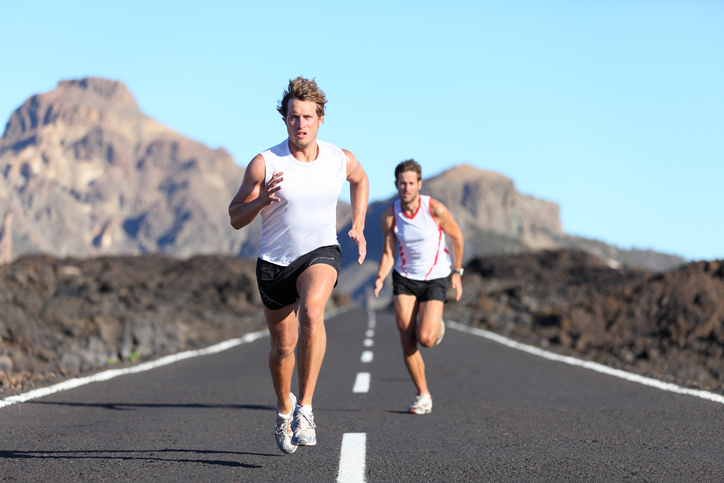Two male runners racing on a road with cliffs and blue sky in background.