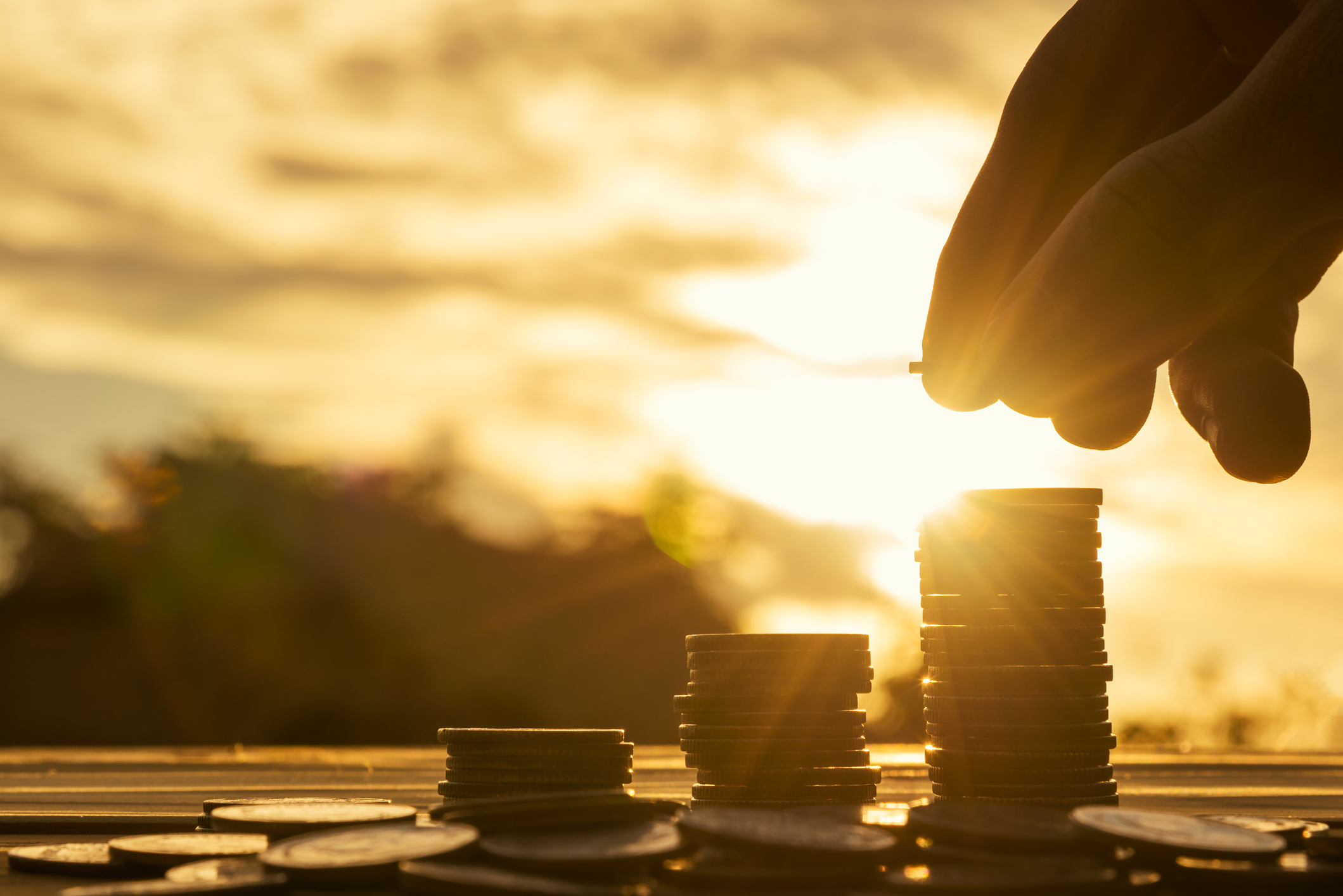 A hand adding a coin to a stack with the sun shining in the background.
