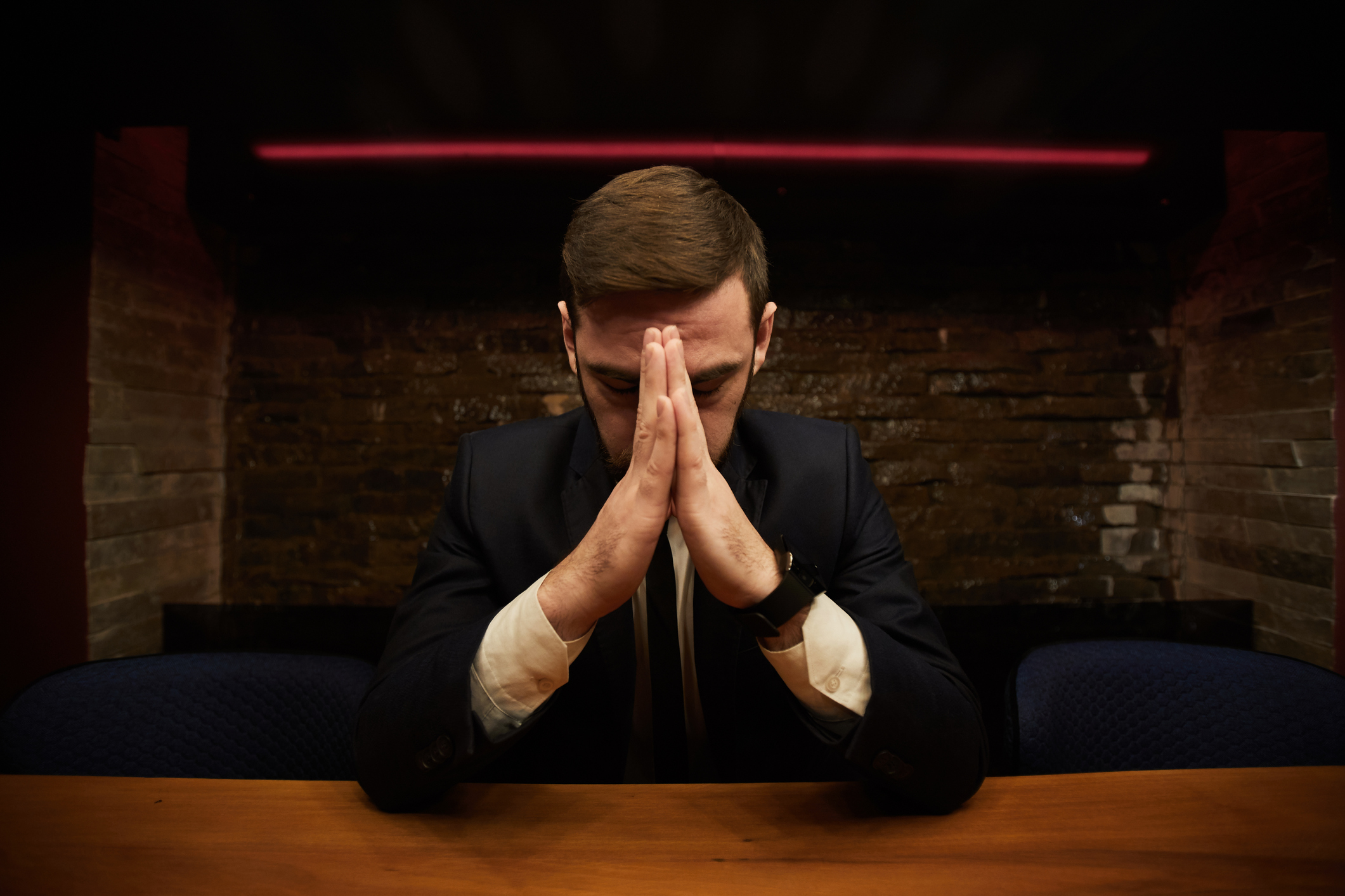 A man sitting at a table with his head bowed and his hands palm to palm in front of his face.