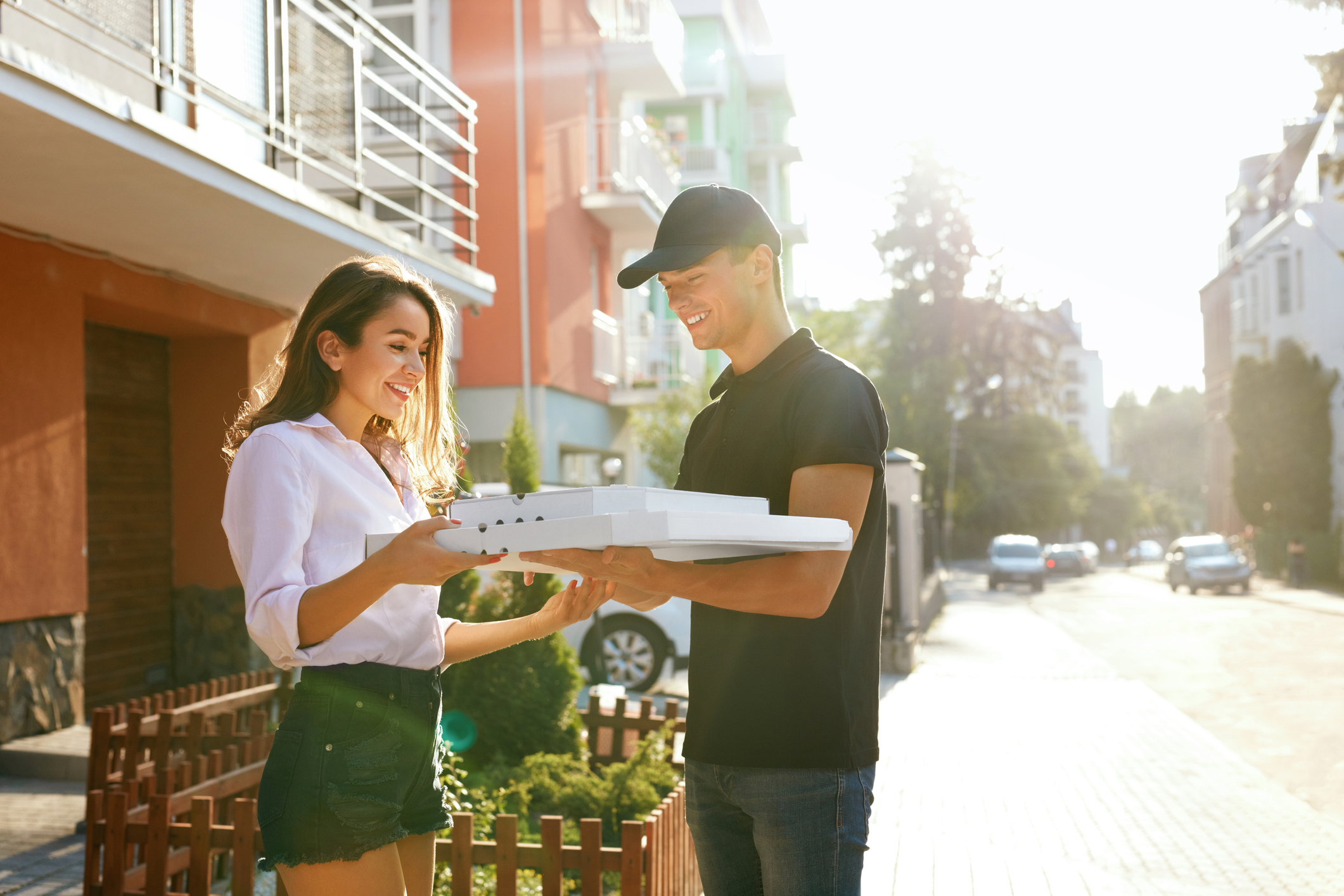 A young woman receives a food delivery, meeting the delivery man on the street and smiling. 