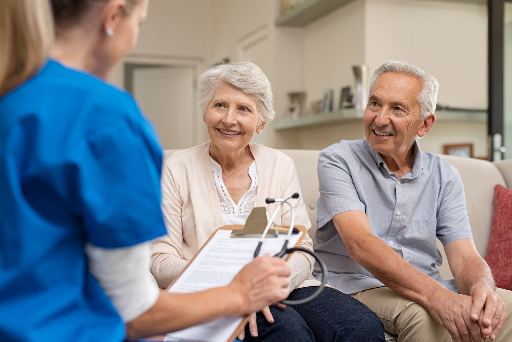 elderly couple sitting on a couch speaking with  healthcare provider in blue scrubs