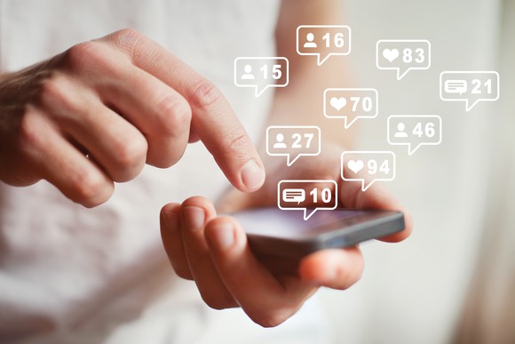 A person holding a smartphone with icons hovering above it, representing users, comments, and likes