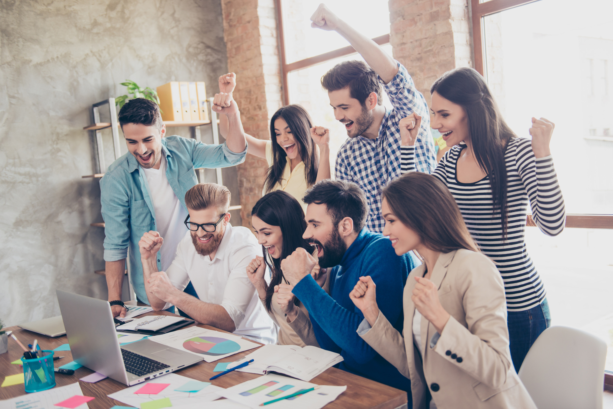 Eight young tech workers gathered around some financial reports and a laptop and cheering.