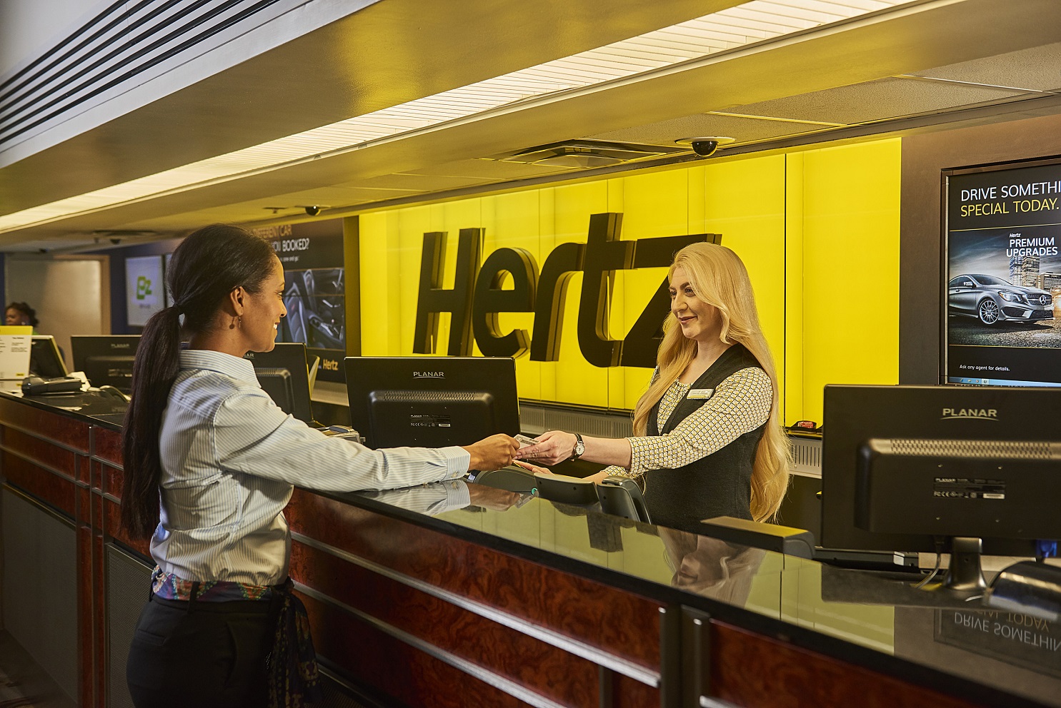 Two people separated by a counter, with the Hertz sign in the background.