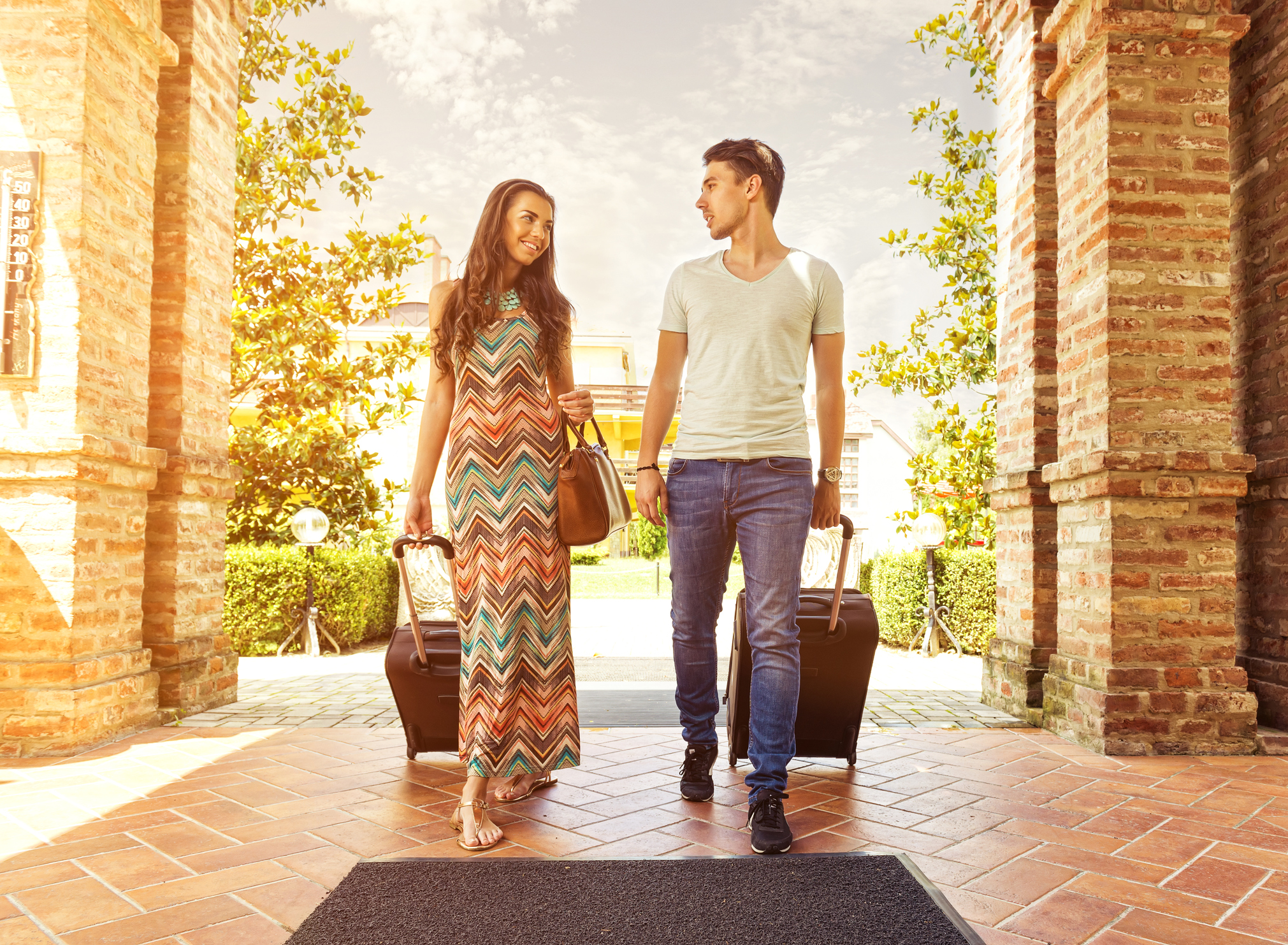 A young couple walking through a hotel portico with wheeled suitcases