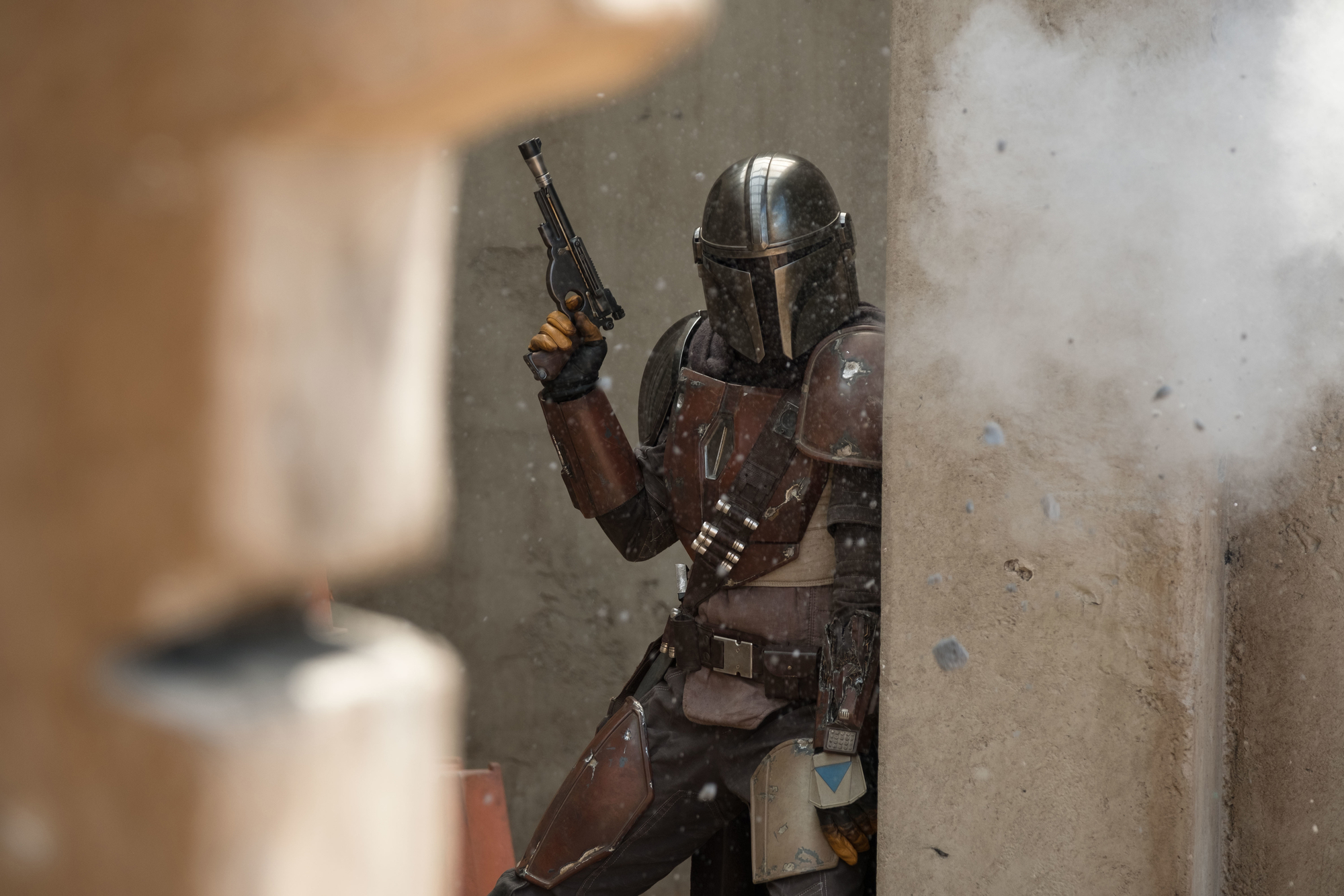 A person in body armor and a helmet holding a gun and taking cover behind a wall.