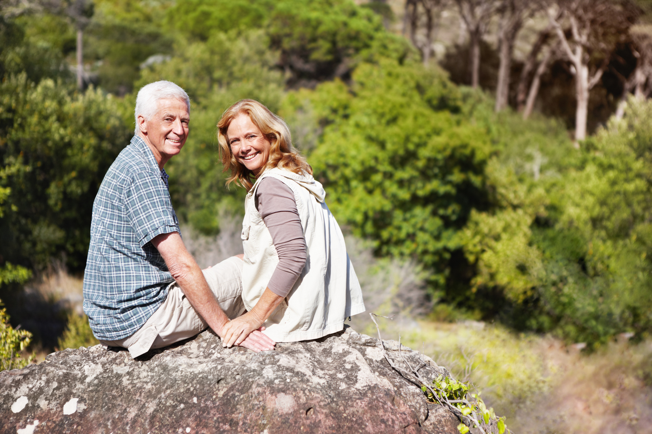 Smiling older man and woman sitting on a rock surrounded by trees.