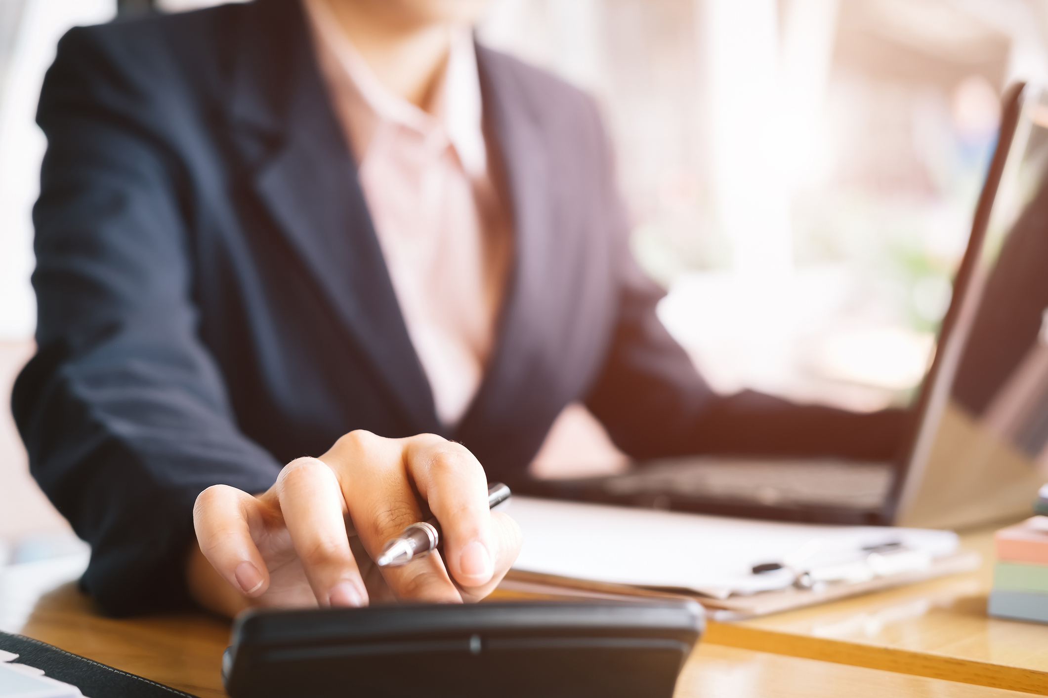 Woman in suit jacket using calculator with laptop nearby.