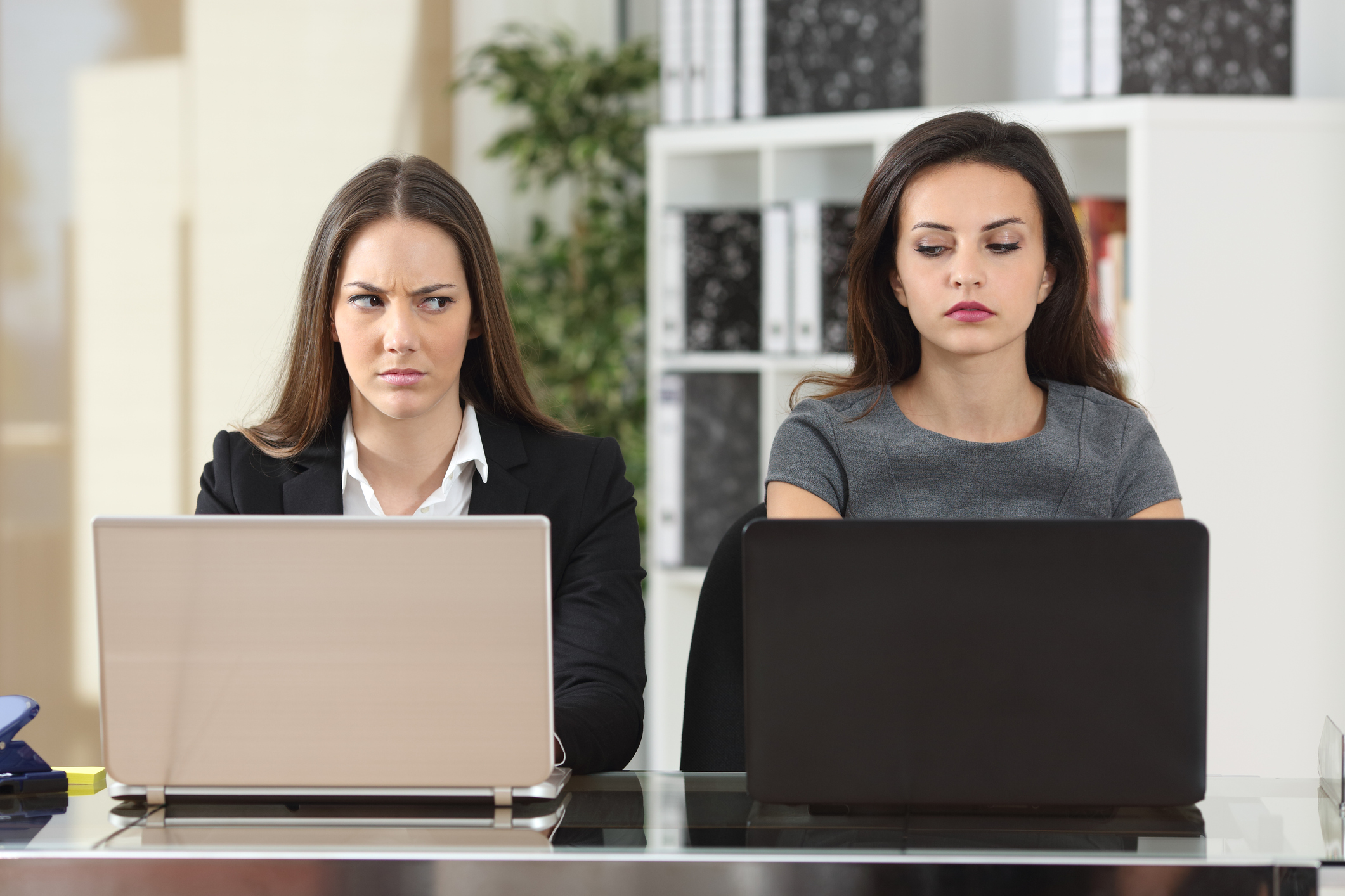 Two women seated next to each other at laptops giving each other dirty looks
