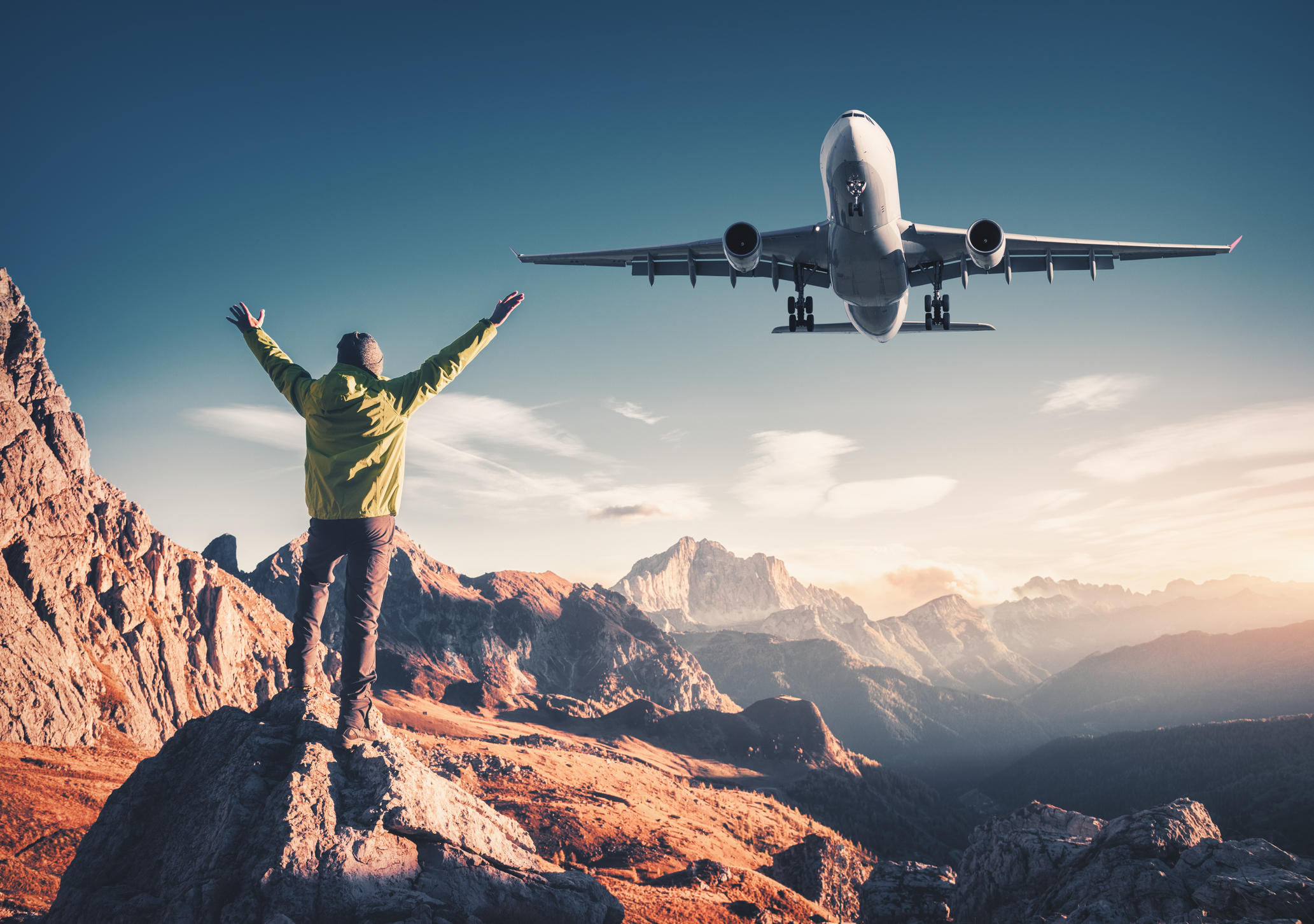 Man surrounded by mountains raising his arms while a plane flies overhead.