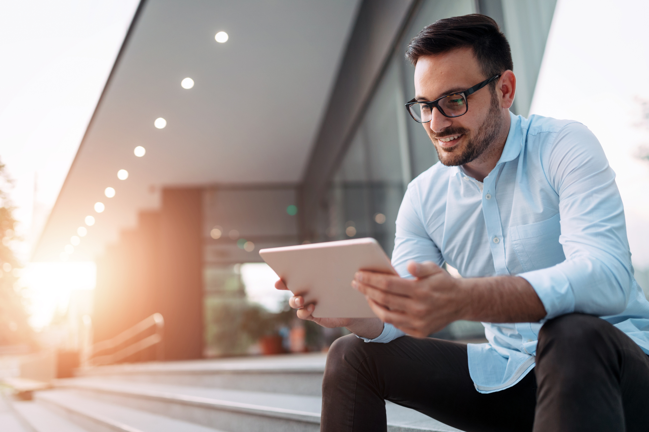 Man sitting on steps in front of office building and looking at his tablet and smiling